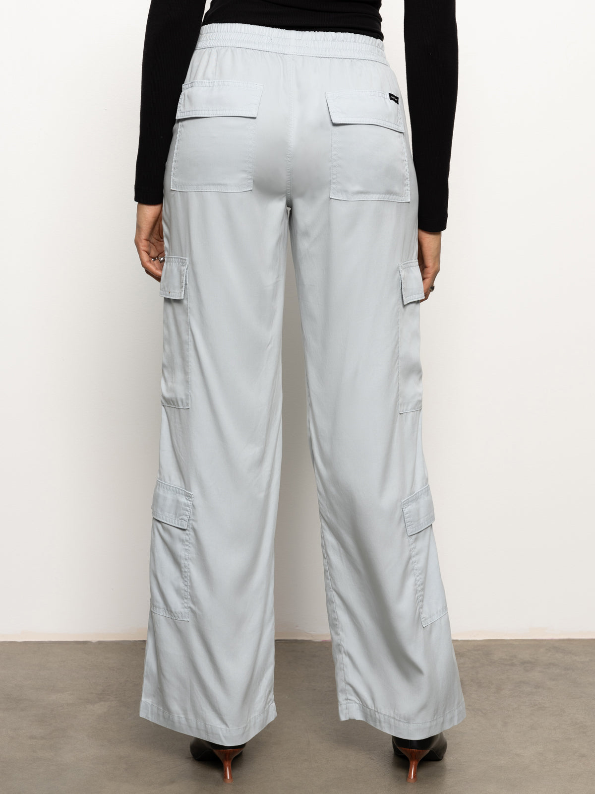 The image features a woman from the back dressed in the "Down to Earth Soft Cargo" pants in Pearl Blue from Sanctuary Clothing, paired with a black long-sleeve top, standing against a plain background.