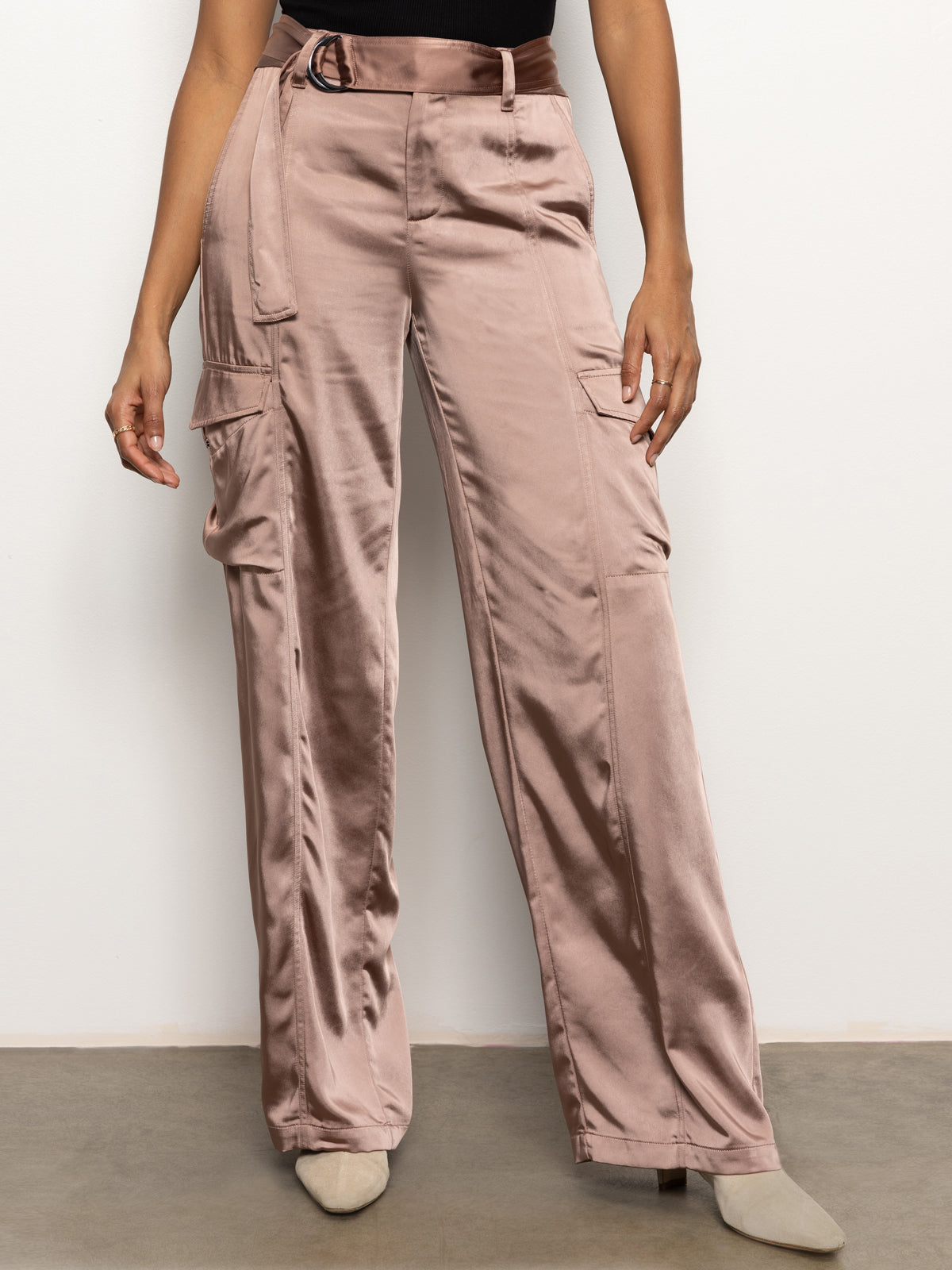 A person is wearing the Classy Wide Leg Cargo Rose Gold pants from Sanctuary Clothing. These satin cargo pants with a shiny finish and large pockets are styled with a black belt and paired with a black top. They are standing against a plain white background.