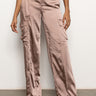 A person is wearing the Classy Wide Leg Cargo Rose Gold pants from Sanctuary Clothing. These satin cargo pants with a shiny finish and large pockets are styled with a black belt and paired with a black top. They are standing against a plain white background.