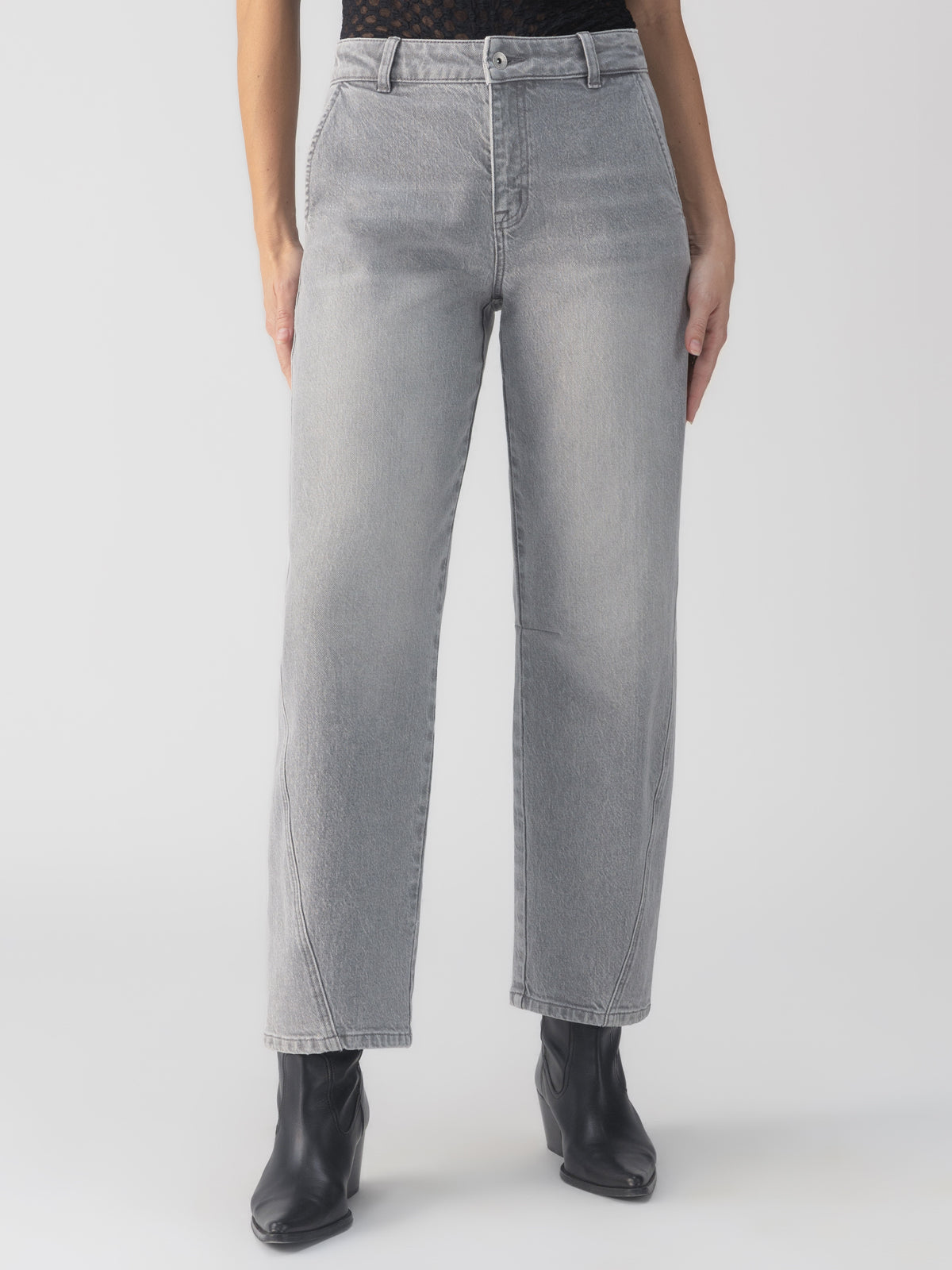 A person wearing the twisted seam jeans barrel grey mist from Sanctuary Clothing, paired with black ankle boots, is standing against a plain white background. The high-waisted jeans feature straight legs, a button and zipper closure, and pockets. The image highlights the lower half of the body.