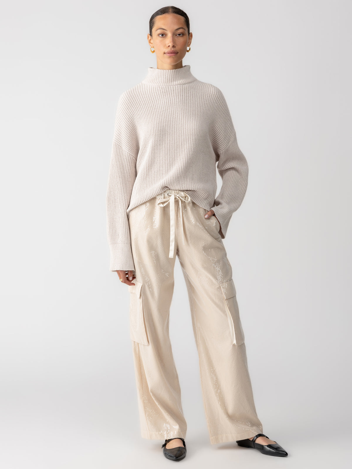 A person stands against a plain background wearing a beige turtleneck sweater, Sanctuary Clothing's Pull On Sequin Cargo Pant in Frosted Almond with a drawstring waist, and black ballet flats. They have their hair pulled back and are wearing gold hoop earrings.