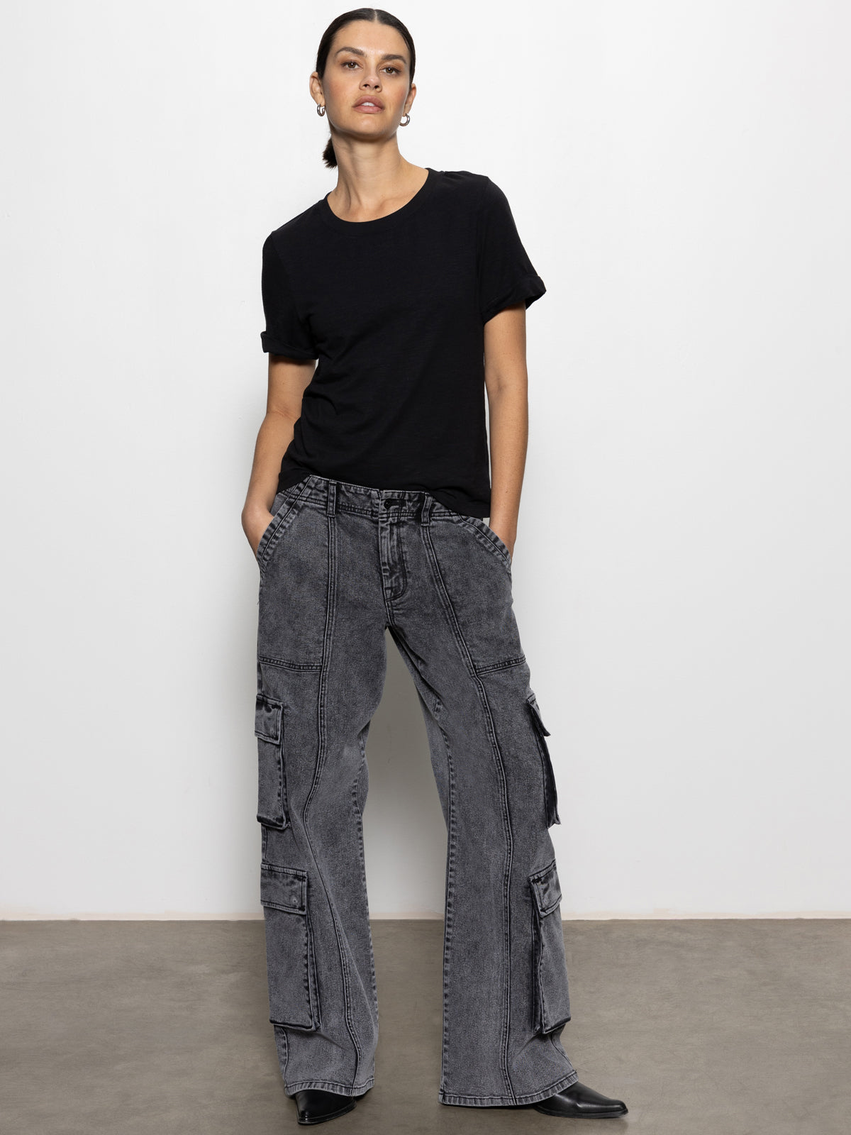 A person stands against a plain white background, wearing the speed denim cargo odyssey from Sanctuary Clothing, styled with multiple pockets. They have their hands in the cargo pants' pockets and are wearing black shoes.