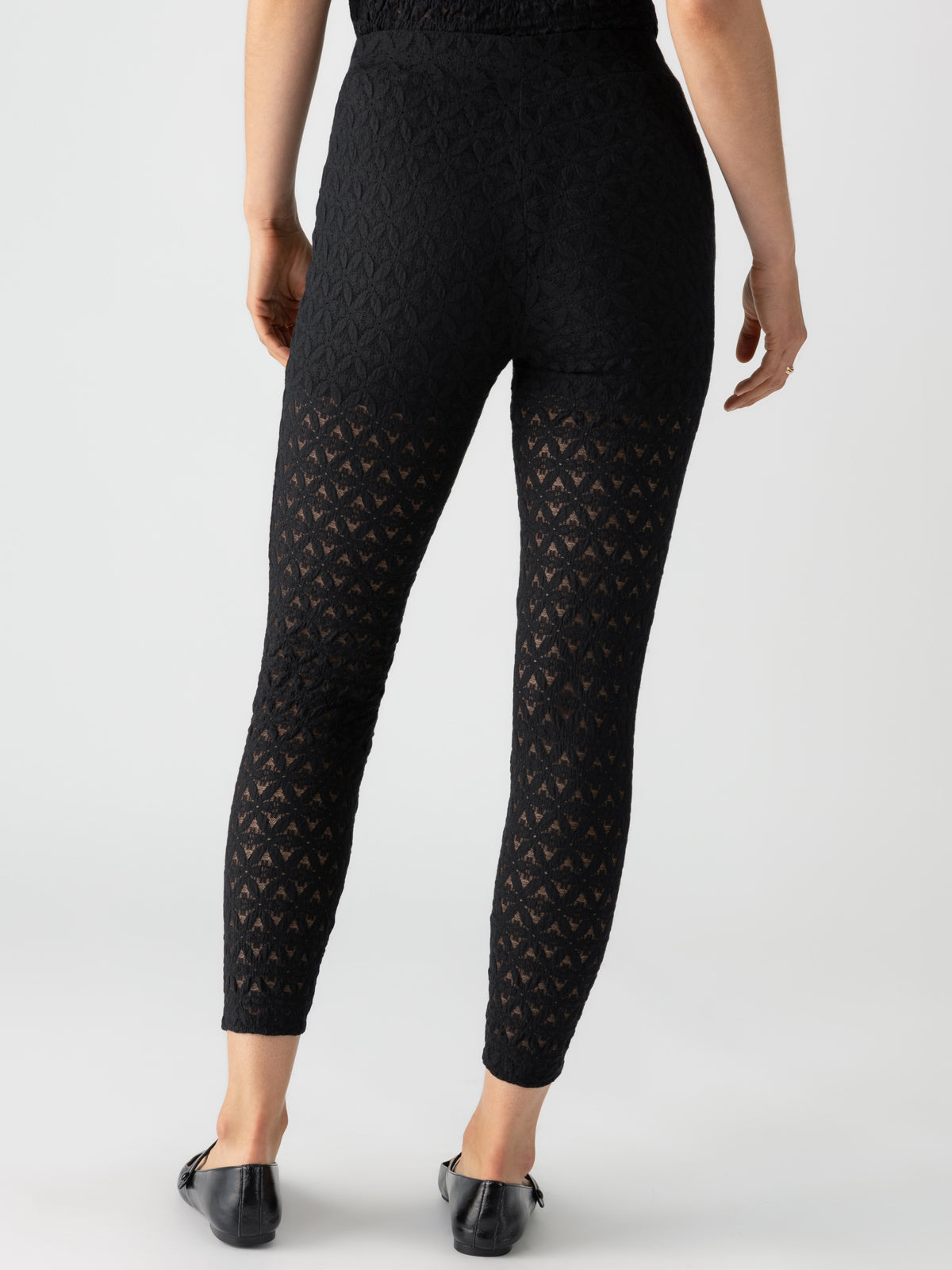 A person wearing the black "Pucker Up Runway Legging" by Sanctuary Clothing paired with black flat shoes is standing with their back facing the camera. The ankle-length leggings feature a sheer patterned design below the knee, all set against a plain white background.