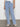 Someone is wearing light blue luna cuffed jeans from Sanctuary Clothing, with button detailing and wide legs, a partially visible white top, and cream-colored crocheted shoes. The background features a plain, light-colored wall and gray floor.