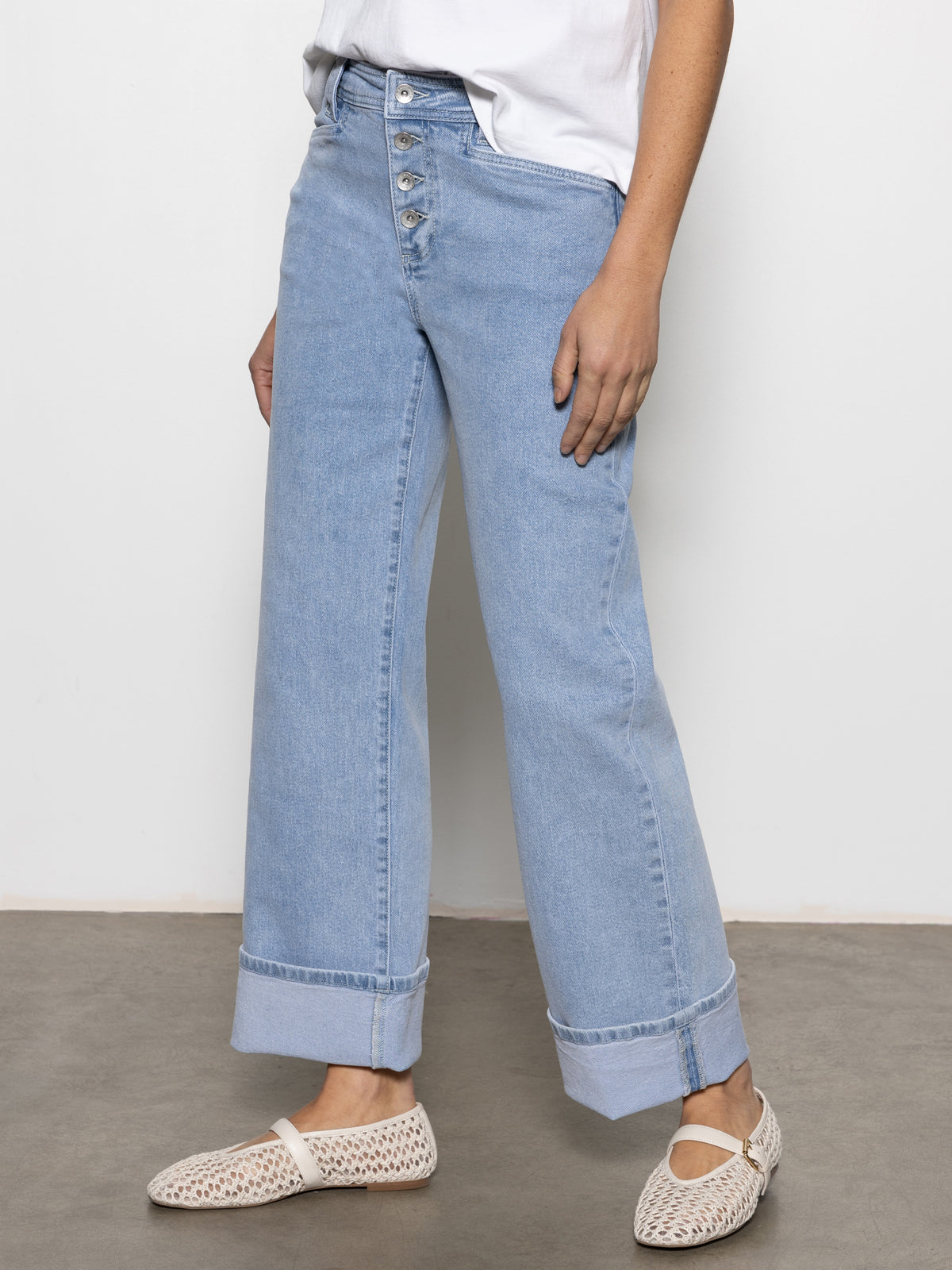 A person wears Sanctuary Clothing's luna cuffed jeans in blue moonlight, paired with a white short-sleeve shirt and white woven slip-on shoes. They stand against a plain white background and concrete floor.