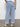 A person is standing with their back to the camera wearing Sanctuary Clothing's "luna cuffed jeans blue moonlight" with large back pockets and a white top tucked in. They pair the jeans with beige woven shoes against a plain light wall and concrete floor.