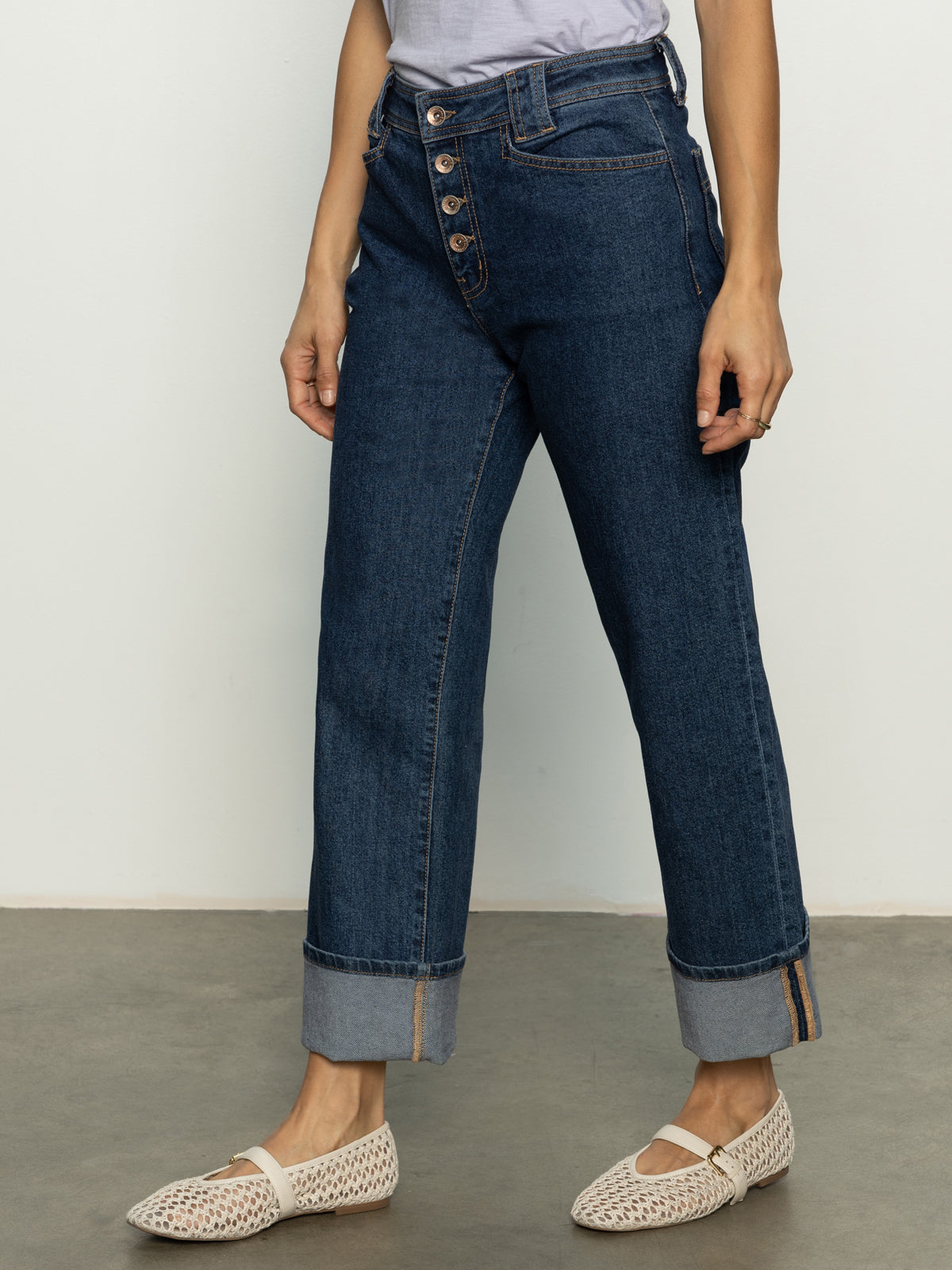 A person wearing the Luna Cuffed Denim Pant Solstice by Sanctuary Clothing stands against a plain background. The high-waisted jeans feature a dark blue hue, rolled cuffs, and a button fly, paired with white woven slip-on shoes.