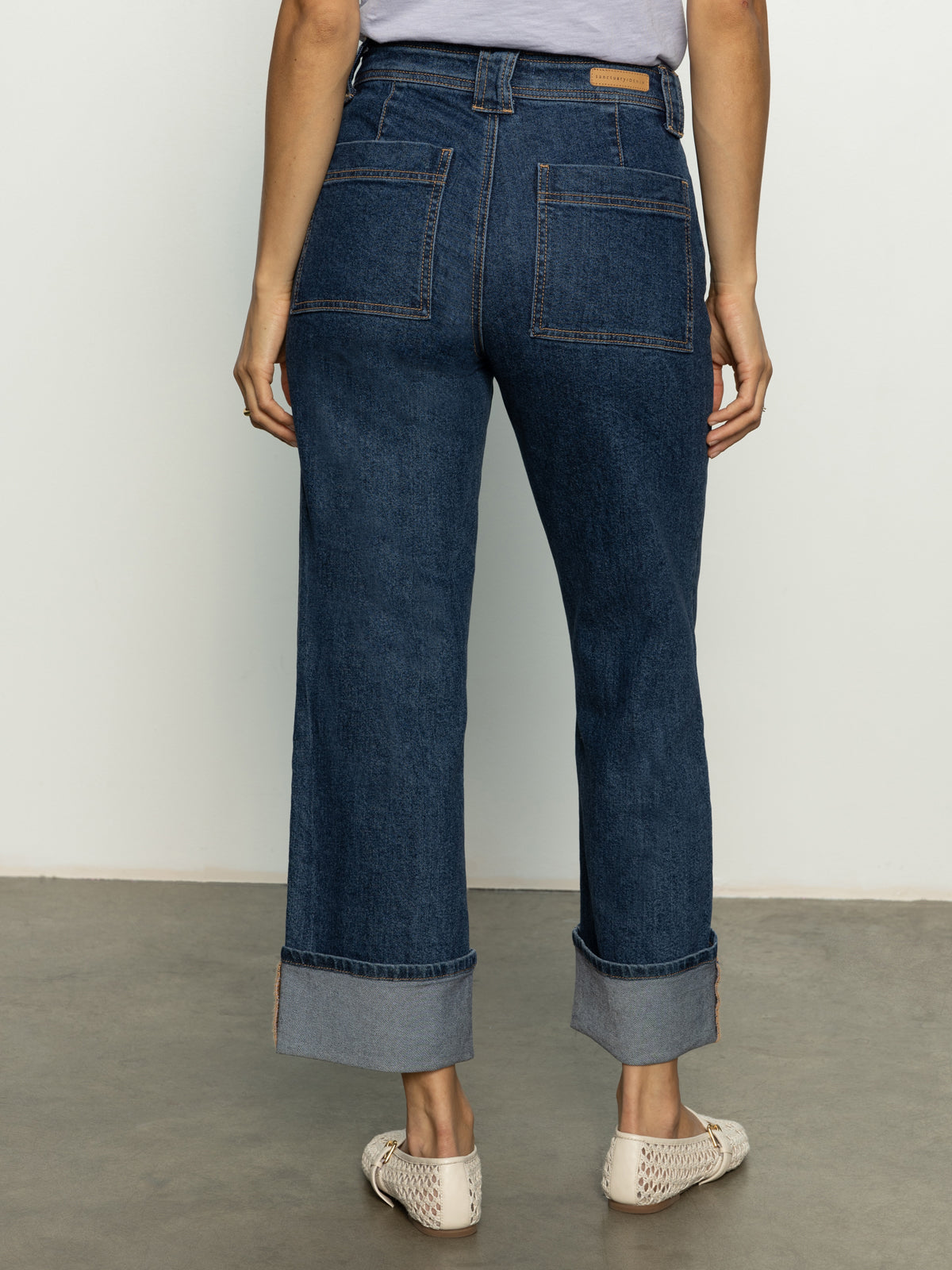 A person is seen from the back wearing Sanctuary Clothing's Luna cuffed denim pant solstice, featuring high-waisted, wide-legged dark denim with large back pockets and rolled cuffs. They pair it with light woven shoes on a smooth gray floor against a plain background.