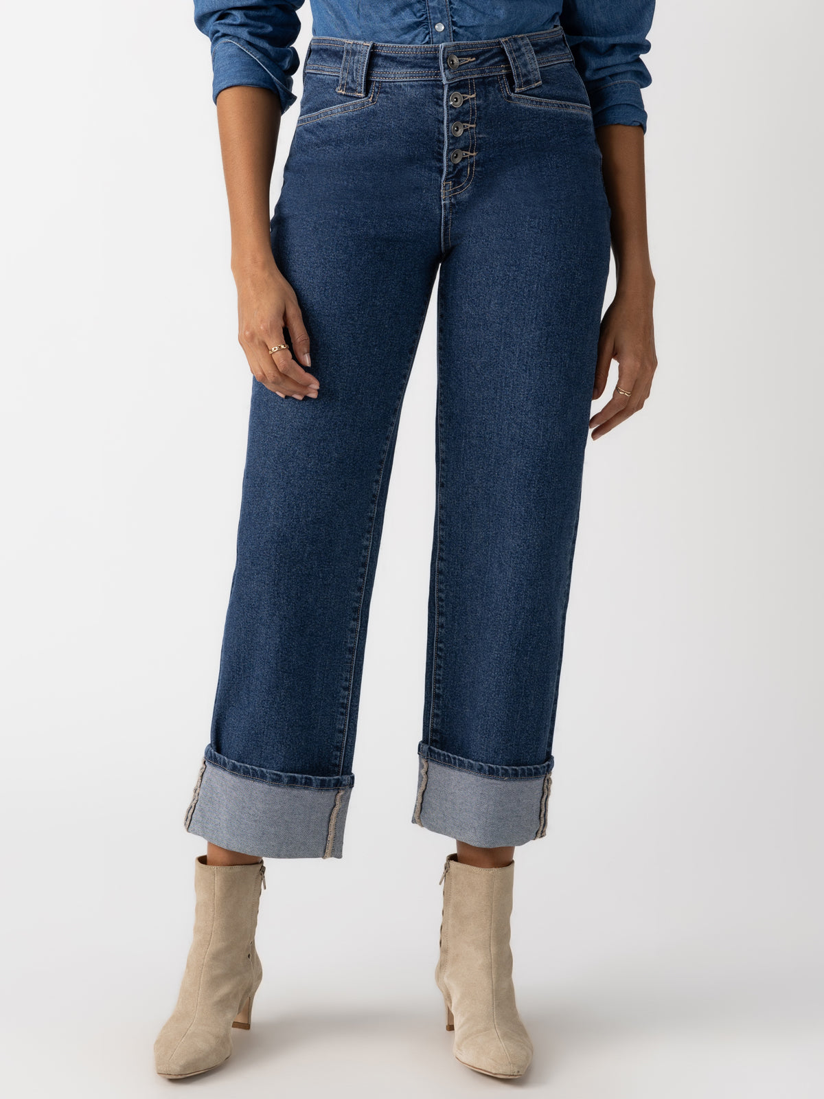 A person wearing the Luna Cuffed Denim Pant Solstice from Sanctuary Clothing, a pair of high-waisted blue jeans with cuffed hems and button details, is paired with an untucked blue shirt and beige ankle boots against a plain white background.