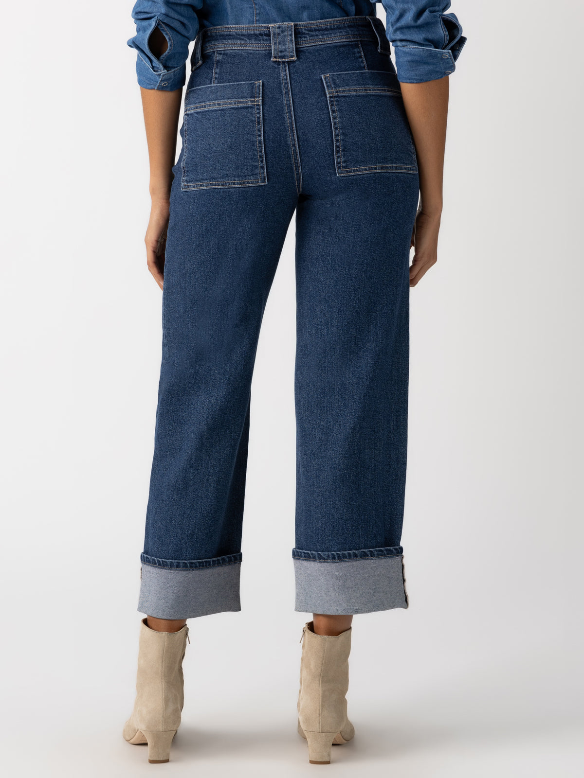 A person is shown from behind wearing the "luna cuffed denim pant solstice" by Sanctuary Clothing. These high-waisted, wide-leg jeans feature large back pockets and are styled with rolled hems. They are paired with light-colored ankle boots and a blue denim jacket.