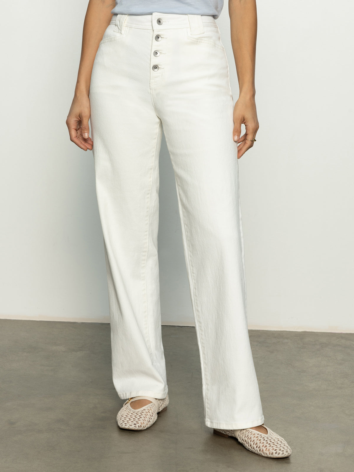 Someone is wearing Sanctuary Clothing's Luna Cuffed Denim Pant in bleached white, featuring a high-waisted design with front buttons, and a blue top. They stand on a smooth gray floor, paired with woven beige slip-on shoes.