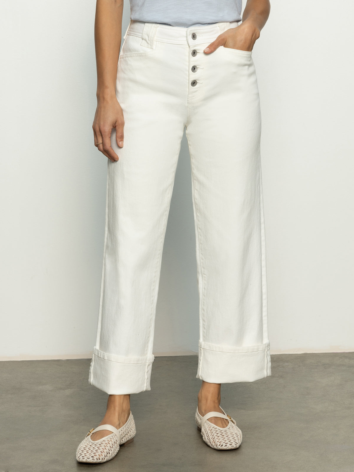 A person wearing Sanctuary Clothing's Luna Cuffed Denim Pant in Bleached White, styled with a light top and woven white flats, stands on a plain floor against a light-colored wall.