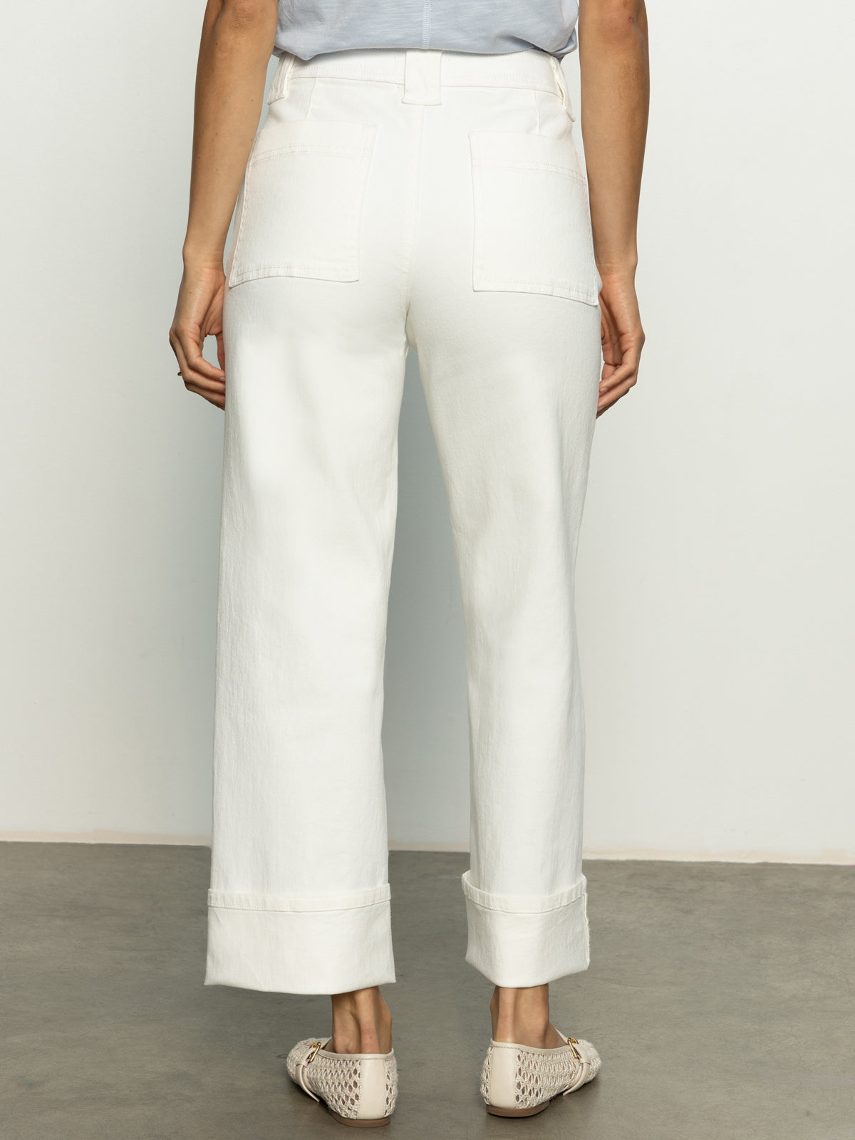 A person stands on a gray surface, wearing Sanctuary Clothing's Luna cuffed denim pant in bleached white. Their top is tucked in, and they sport woven slip-on shoes against a plain white background.