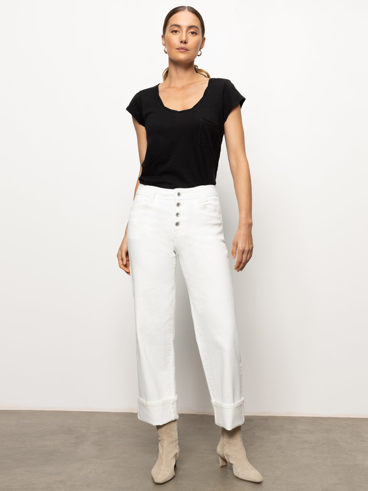 A person stands against a plain white background, wearing a black t-shirt and the Luna Cuffed Denim Pant in bleached white from Sanctuary Clothing. They are styled with beige ankle boots and hoop earrings, and their hair is tied back.