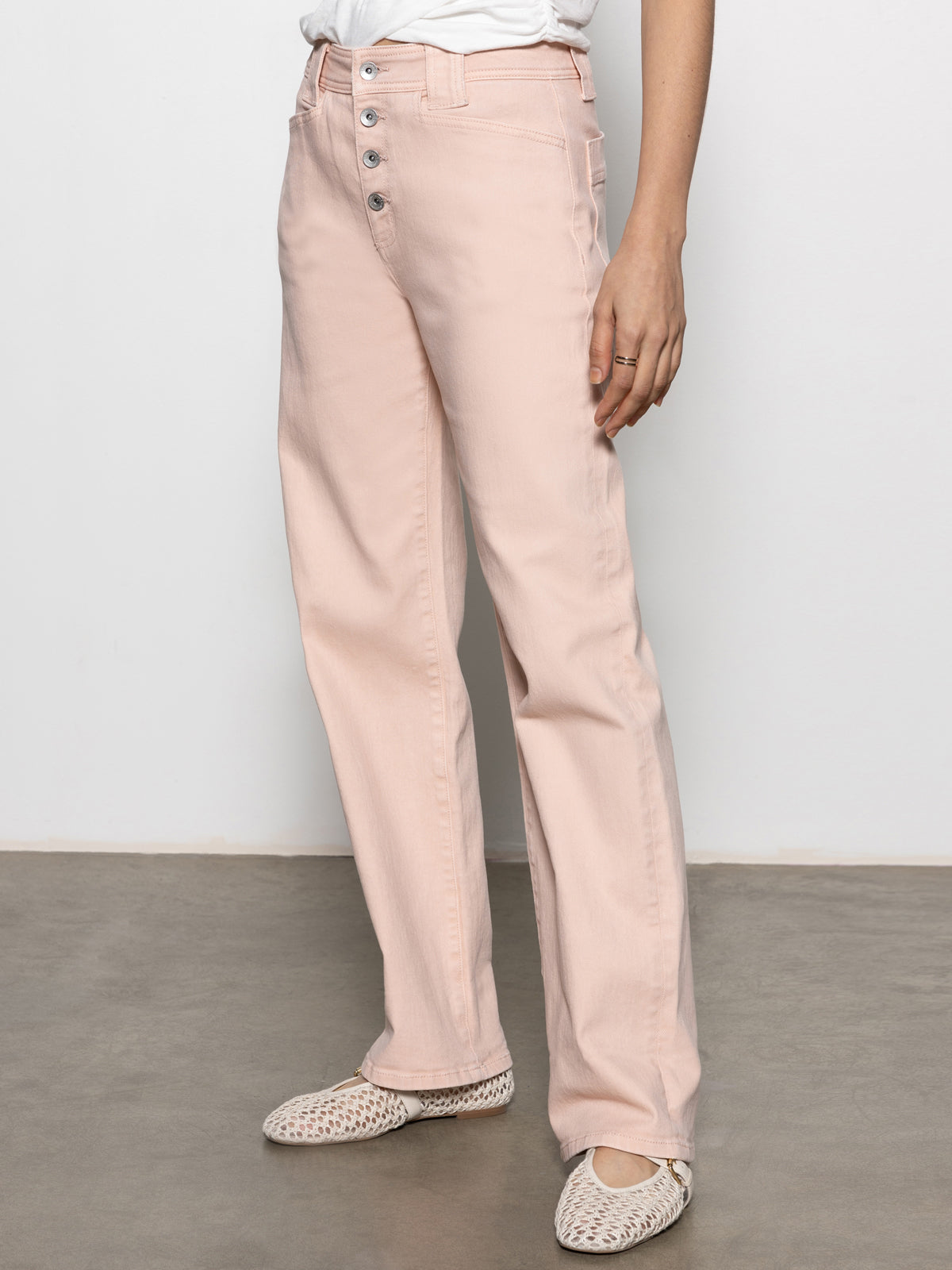 A person wearing Sanctuary Clothing's Luna Cuffed Jeans in Earth Pink, featuring a high-waist and four buttons, paired with a white top stands against a light gray wall. They complete the look with woven white flats.