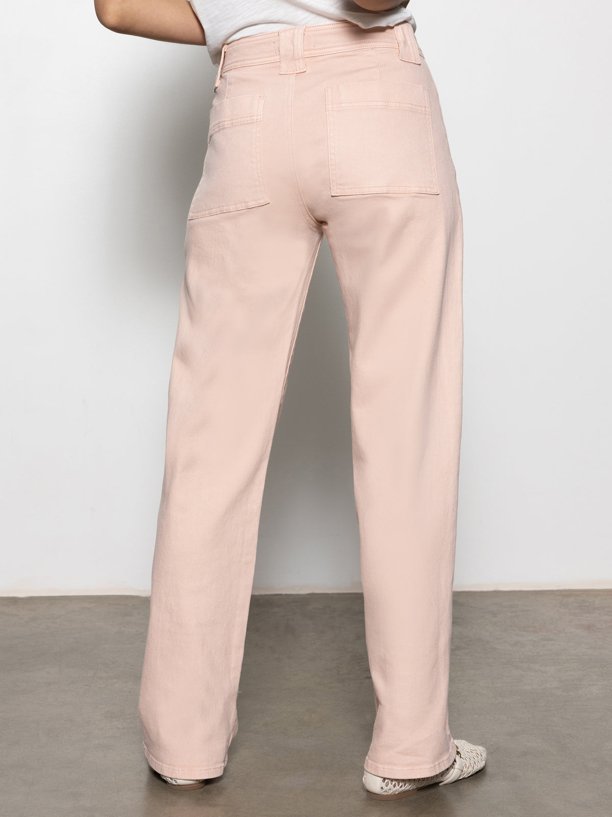 A person stands against a plain wall on a concrete floor wearing the Sanctuary Clothing "Luna" cuffed jeans in earth pink. They pair these high-waisted, wide-leg pants featuring rear patch pockets with a white top and patterned shoes.