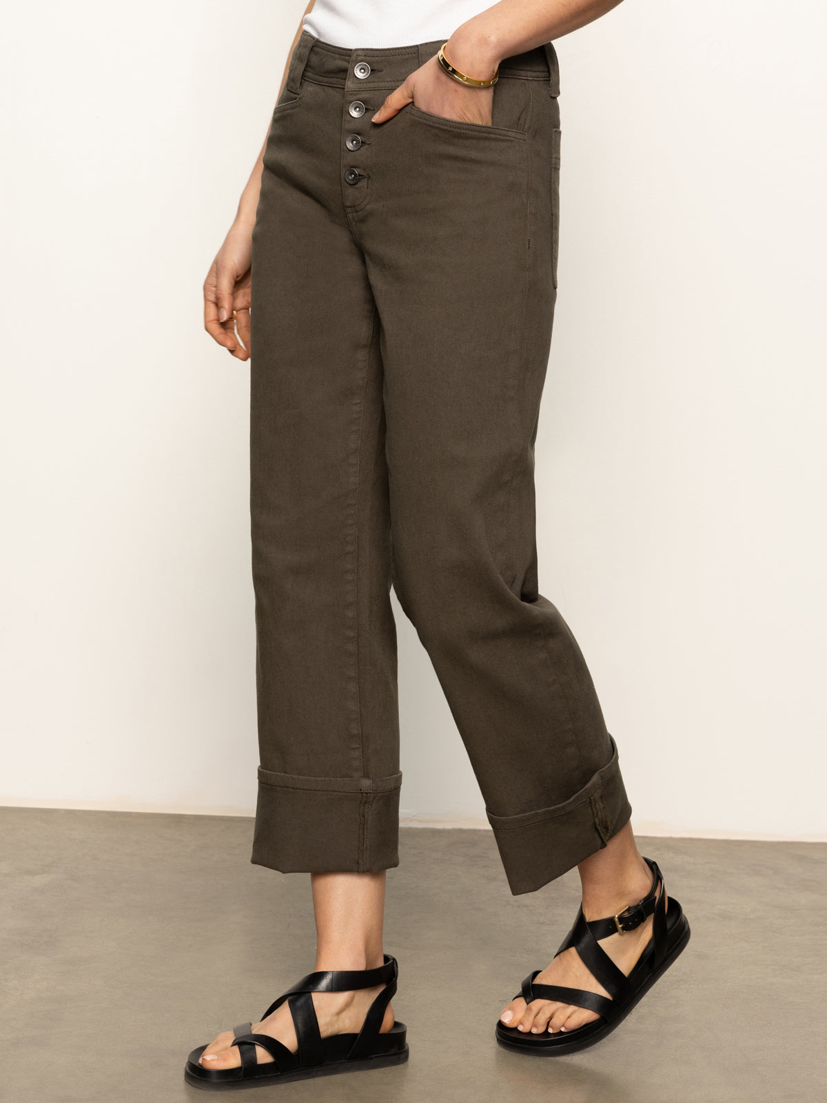 The individual is wearing the "Luna Cuffed Jeans Kalamata" by Sanctuary Clothing, featuring cropped dark olive pants with button front and pocket detail, paired with black strappy sandals. They stand on a neutral floor against a light background.