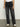 Someone in a Sanctuary Clothing luna cuffed denim pant dark smoke and a white shirt stands on a concrete floor, paired with white sneakers featuring black soles. The plain background is light-colored.