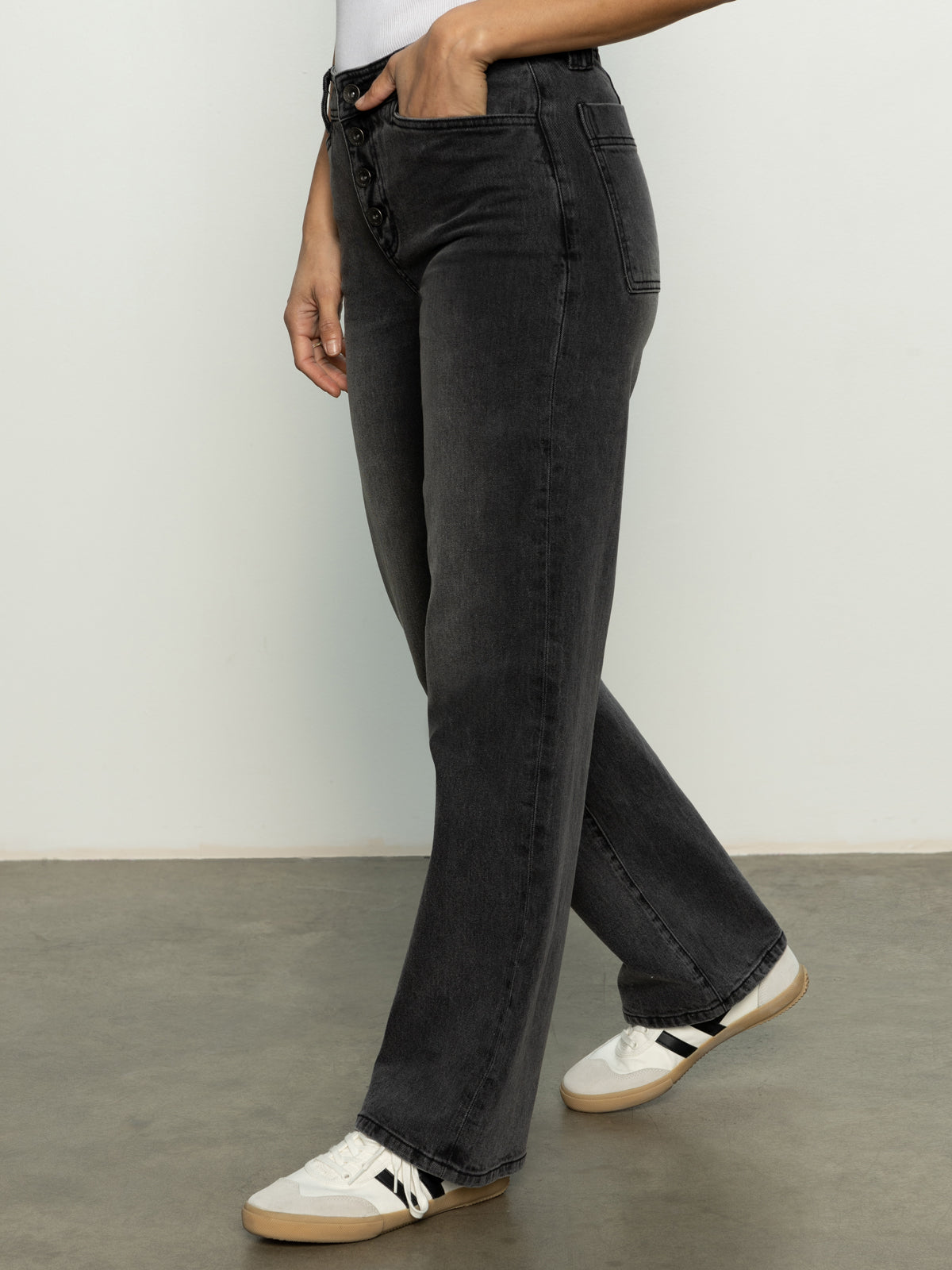 Person wearing Sanctuary Clothing's Luna Cuffed Denim Pant in Dark Smoke paired with white sneakers featuring black and tan details, set against a light-colored wall and gray floor, with one hand in their pocket.