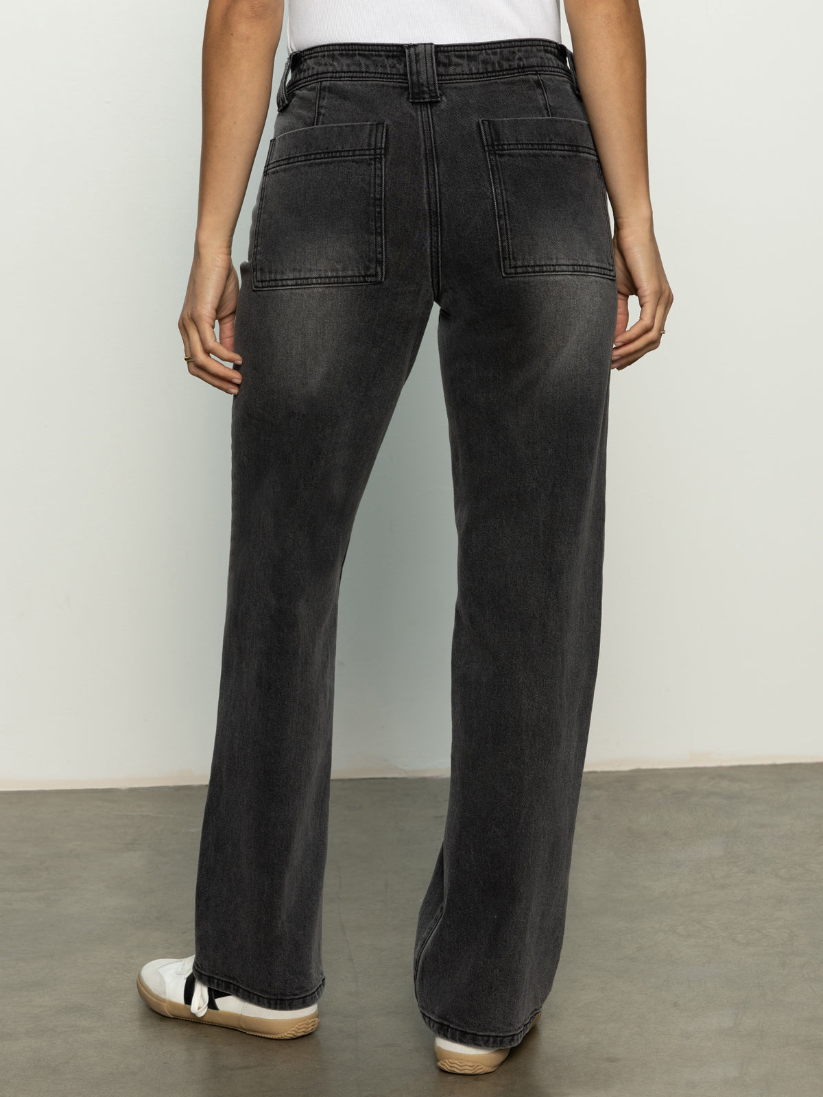 Rear view of a person wearing Luna Cuffed Denim Pant in Dark Smoke by Sanctuary Clothing, featuring two large back pockets. They pair the jeans with white sneakers and a white top against a plain light wall and floor backdrop.