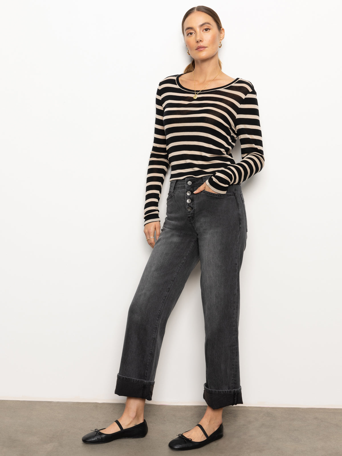 A person stands against a light wall, dressed in a black and beige striped long-sleeve top paired with Sanctuary Clothing's "luna cuffed denim pant" in dark smoke. They are wearing black flats, with their arms relaxed by their sides.