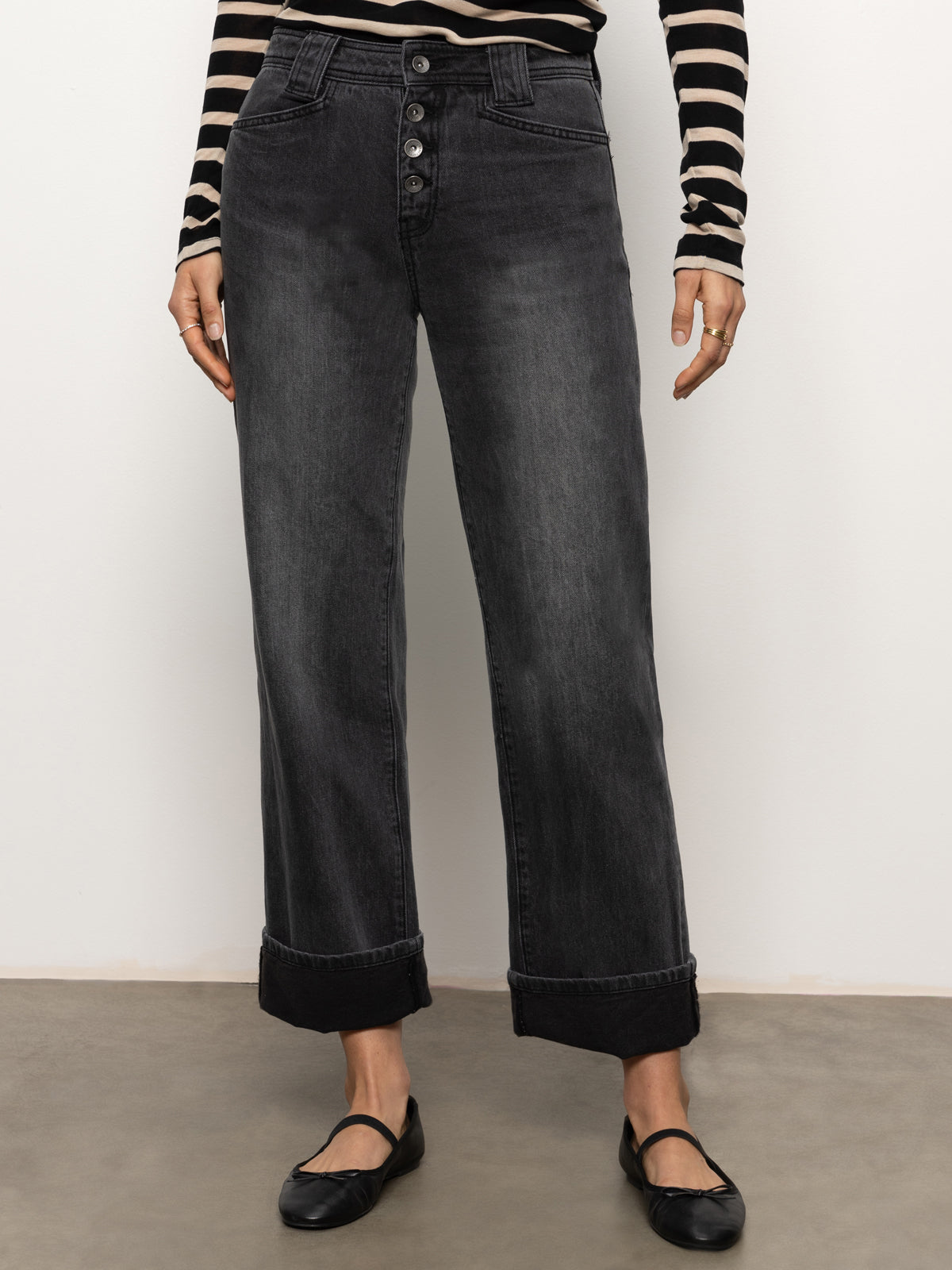 A person is standing against a neutral background, wearing the Luna Cuffed Denim Pant in Dark Smoke by Sanctuary Clothing. They have paired it with a black and white striped long-sleeve shirt and black flats.