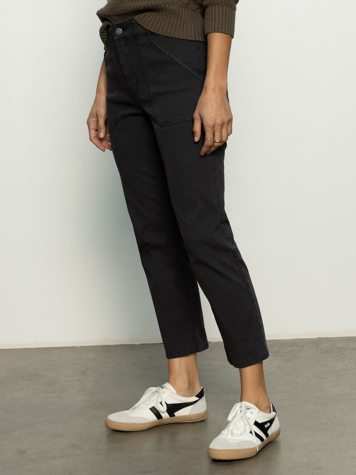 A person wears Sanctuary Clothing's sculpted Hayden straight crop pant in black and white sneakers, standing on a grey floor. They have a green sweater on. Only their lower half is visible, with their right hand resting on their thigh.