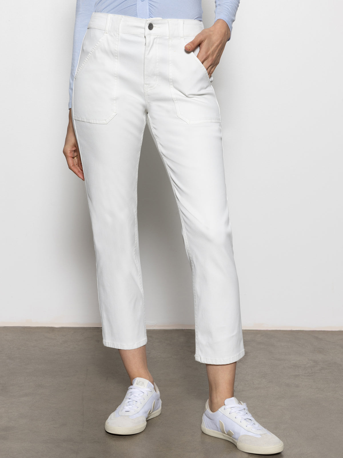 A person is wearing Sanctuary Clothing's sculpted Hayden straight crop pants in brilliant white, paired with white sneakers featuring pale yellow accents. They stand against a plain background, one hand in their pocket, and they're also in a light blue top.