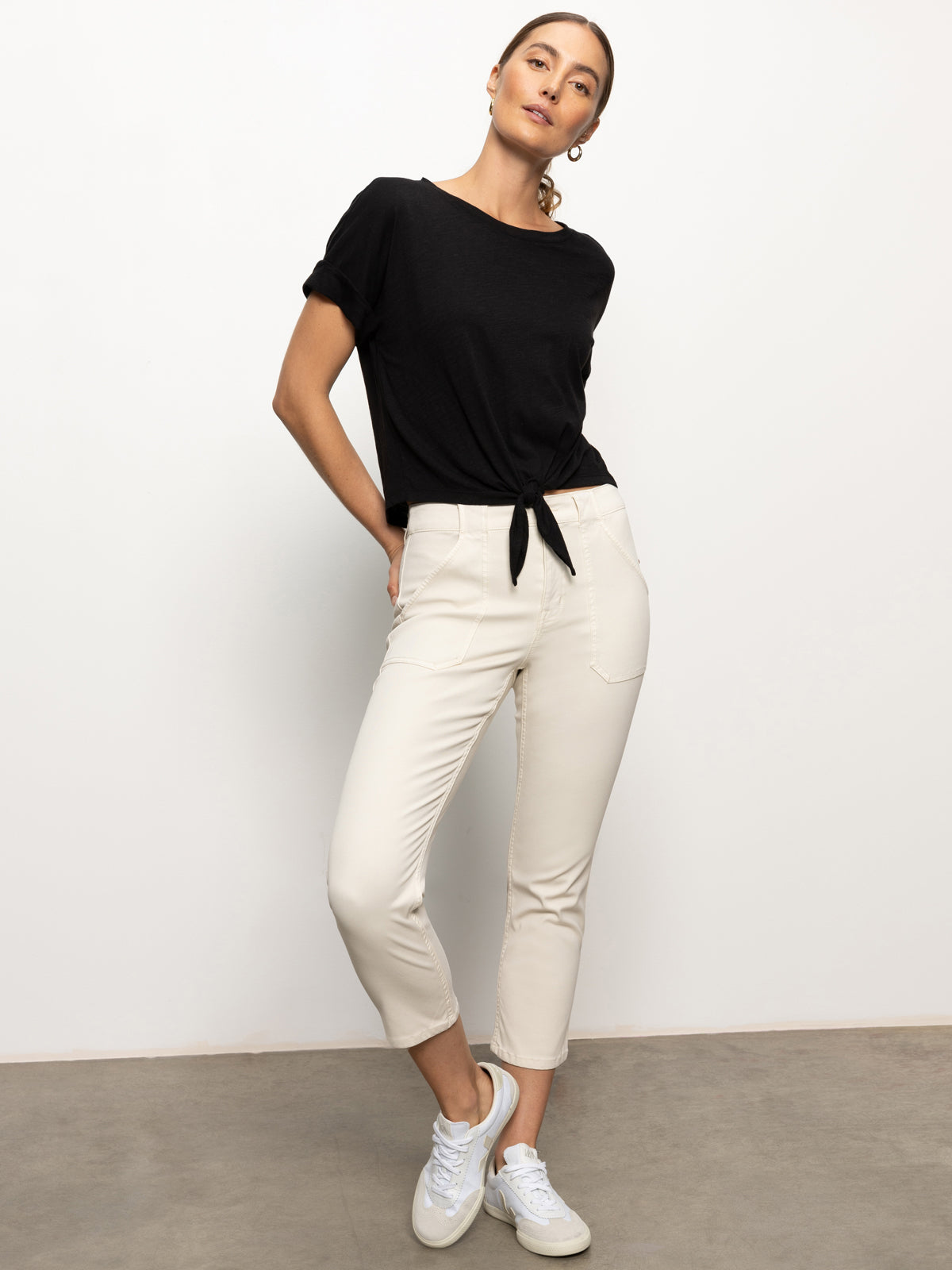 Against a plain white background, the person is wearing a black knotted t-shirt paired with Sculpted Hayden Straight Crop pants in Light Oat by Sanctuary Clothing and white sneakers. Their hands rest in their pockets, their head slightly tilted, and their hair tied back.