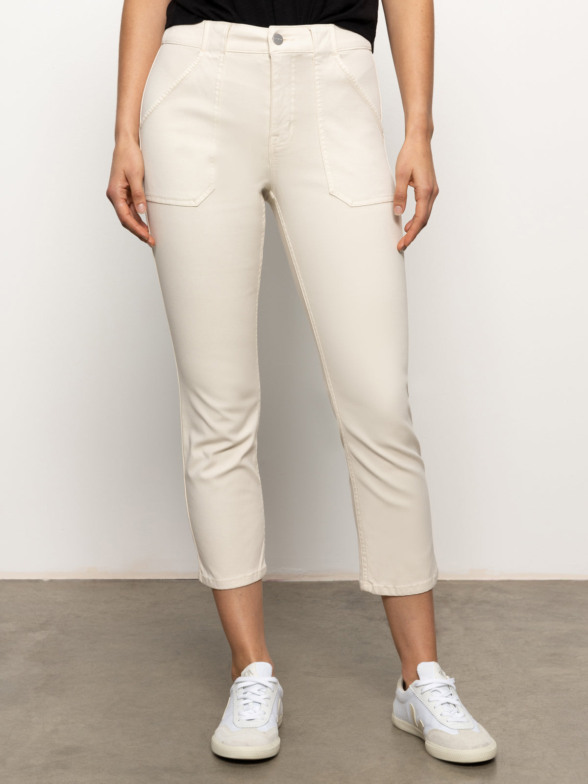 A person is standing on a grey floor wearing Sanctuary Clothing’s sculpted hayden straight crop pants in light oat paired with white sneakers. The pants feature front seam details and large pockets, while the top is black but only partially visible. The background is plain white.