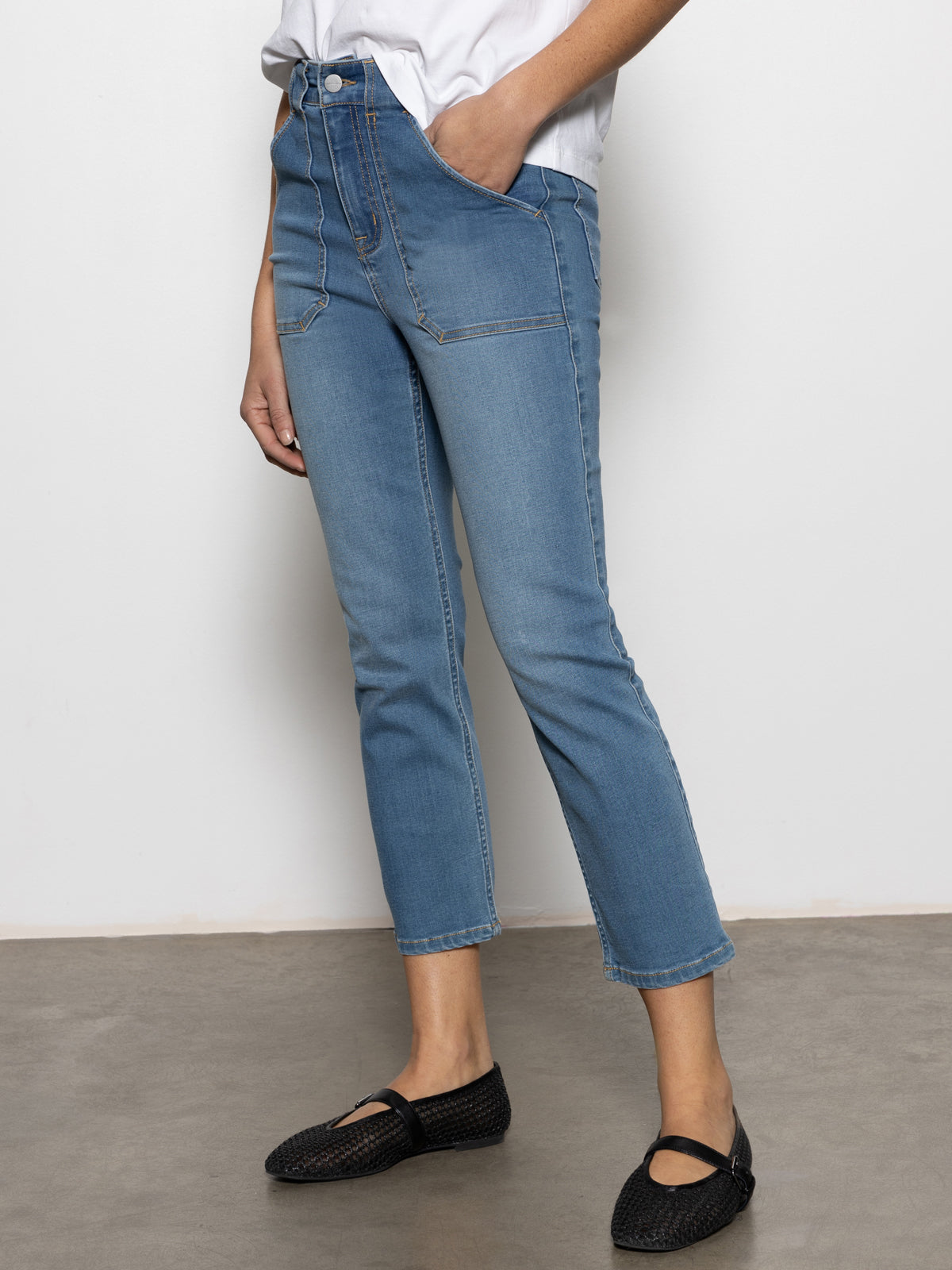 A person wearing the Sanctuary Clothing sculpted Hayden straight crop jeans in 'best in show,' styled with a white top, stands on a gray floor. They complete their look with black woven shoes and have one hand tucked into a pocket of the slightly cropped, high-waisted jeans.