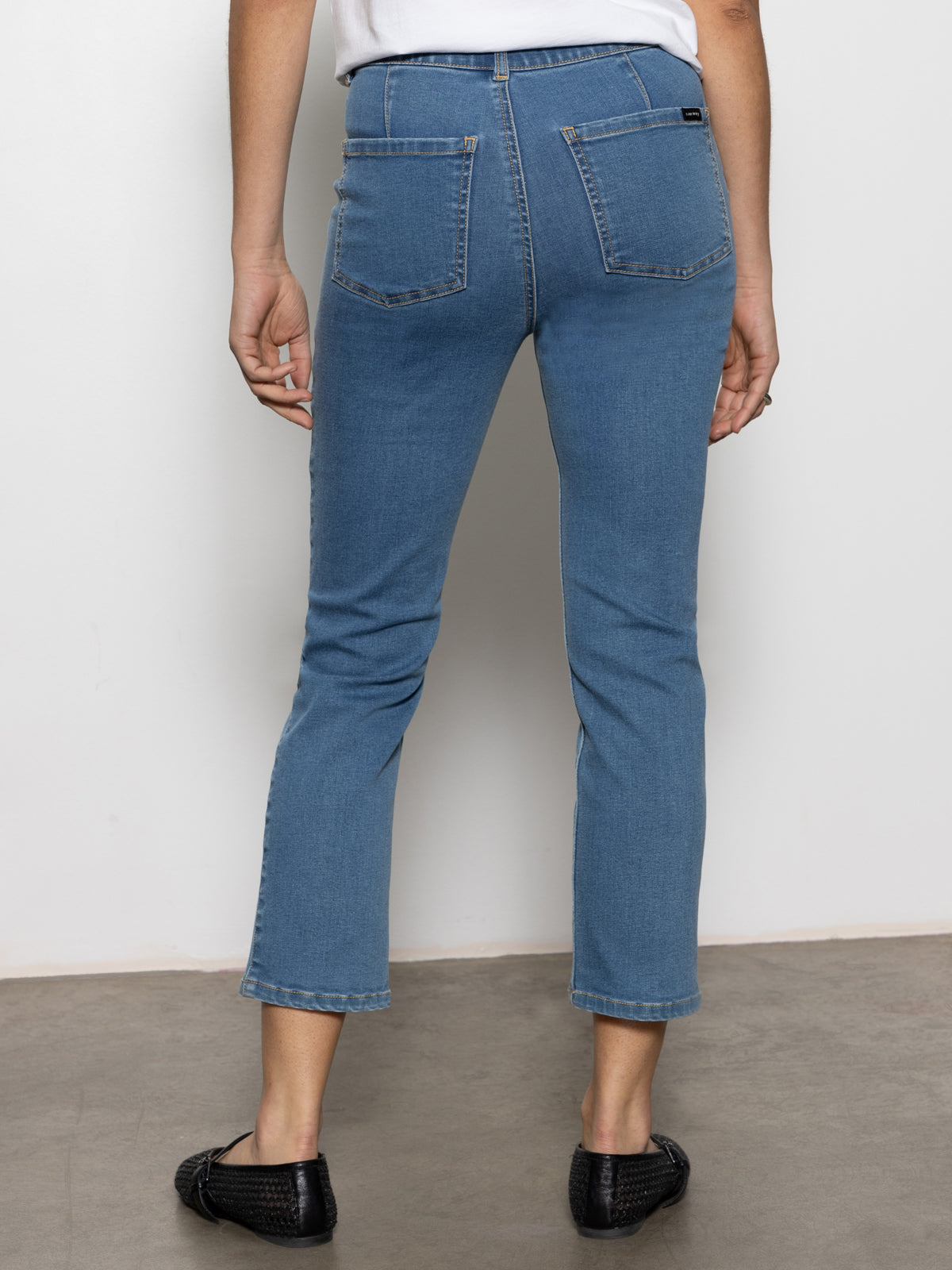 A person is standing on a concrete floor, facing away from the camera. They are wearing the "Sculpted Hayden Straight Crop Best in Show" blue jeans by Sanctuary Clothing, paired with a white shirt and black shoes. The background features a plain white wall.