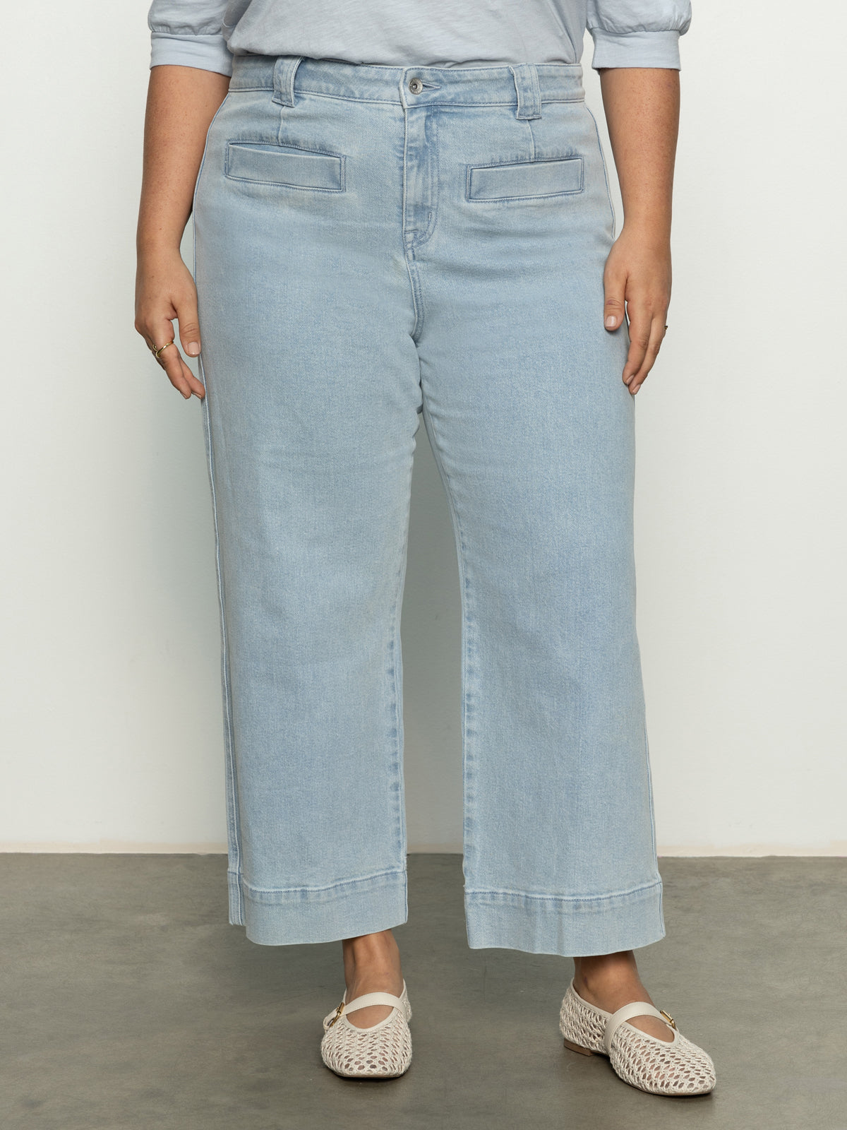 A person in Sanctuary Clothing's voyage crop pant winners circle inclusive collection and a light blue shirt stands on a concrete floor, hands resting at their sides. They wear white woven slip-on shoes, with an off-white wall in the background.