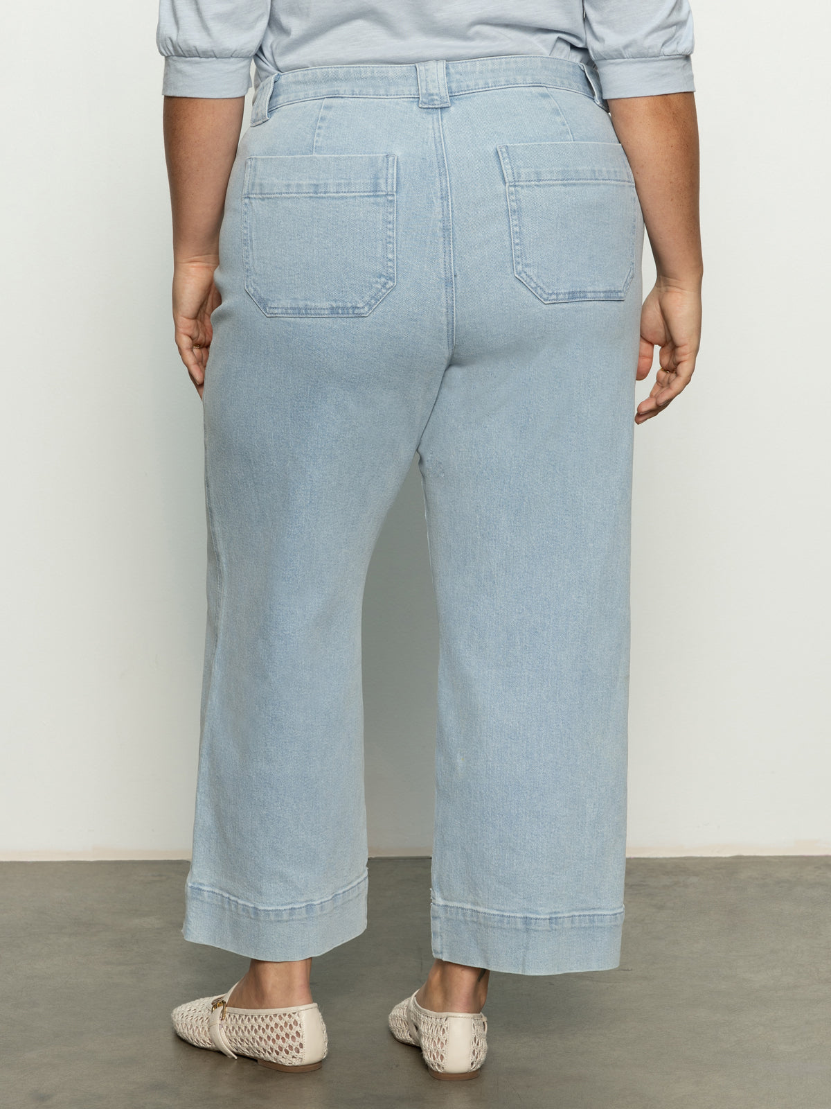 Rear view of a person wearing Voyage Crop Pant from the Winners Circle Inclusive Collection by Sanctuary Clothing, a gray long-sleeve shirt, and beige woven flats. They stand on a gray floor against a white background.