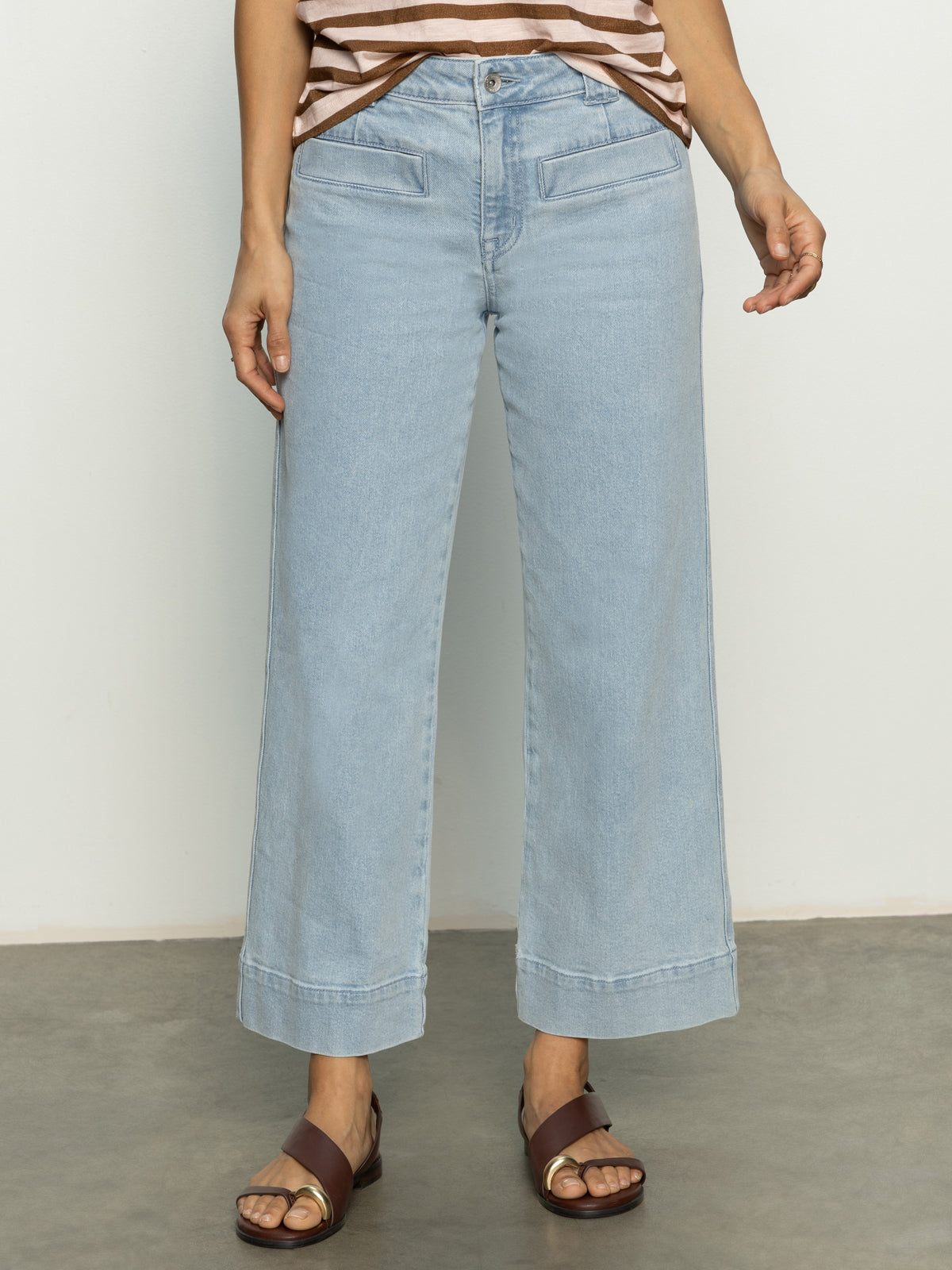 Someone is wearing Sanctuary Clothing's voyage crop pant winners circle, light blue wide-leg jeans with front pockets and ankle-length. They're paired with a brown and white striped sleeveless top and brown sandals, standing on a concrete floor.