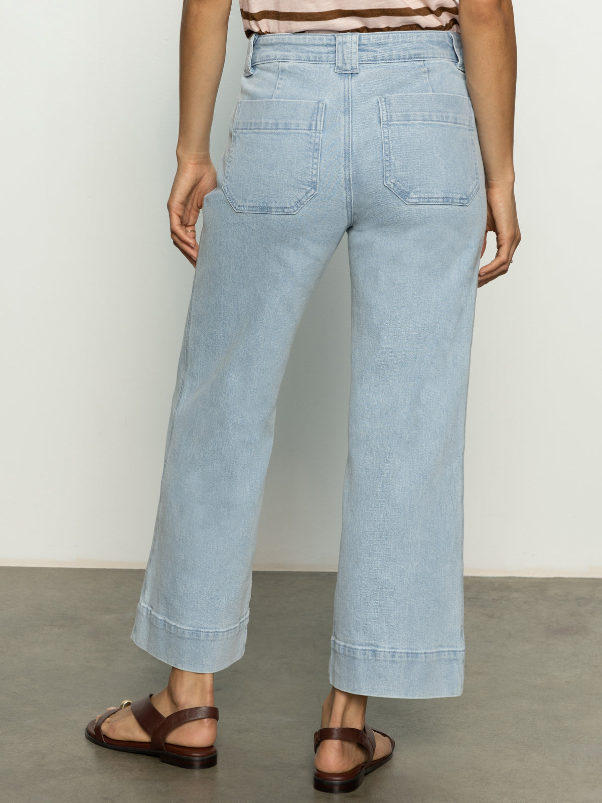 A person wearing Sanctuary Clothing's "voyage crop pant winners circle" in light blue, high-waisted, wide-leg style with two back pockets, paired with brown sandals. They stand on a gray floor against a plain white background.