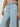 A person wearing Voyage Crop Pant Winners Circle by Sanctuary Clothing stands against a plain background. The light blue high-waisted jeans feature prominent seams and front pockets. Their hands, sporting a ring on one finger, rest at their sides.