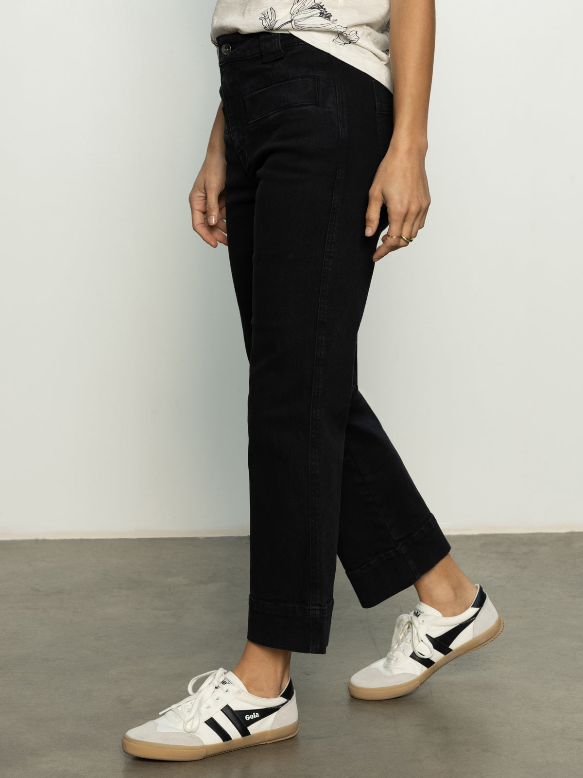 A person is standing on a concrete floor wearing a pair of Sanctuary Clothing's Voyage Crop Pants in black, paired with white sneakers featuring black stripes and a light-colored shirt. Only the lower half of their body is visible.