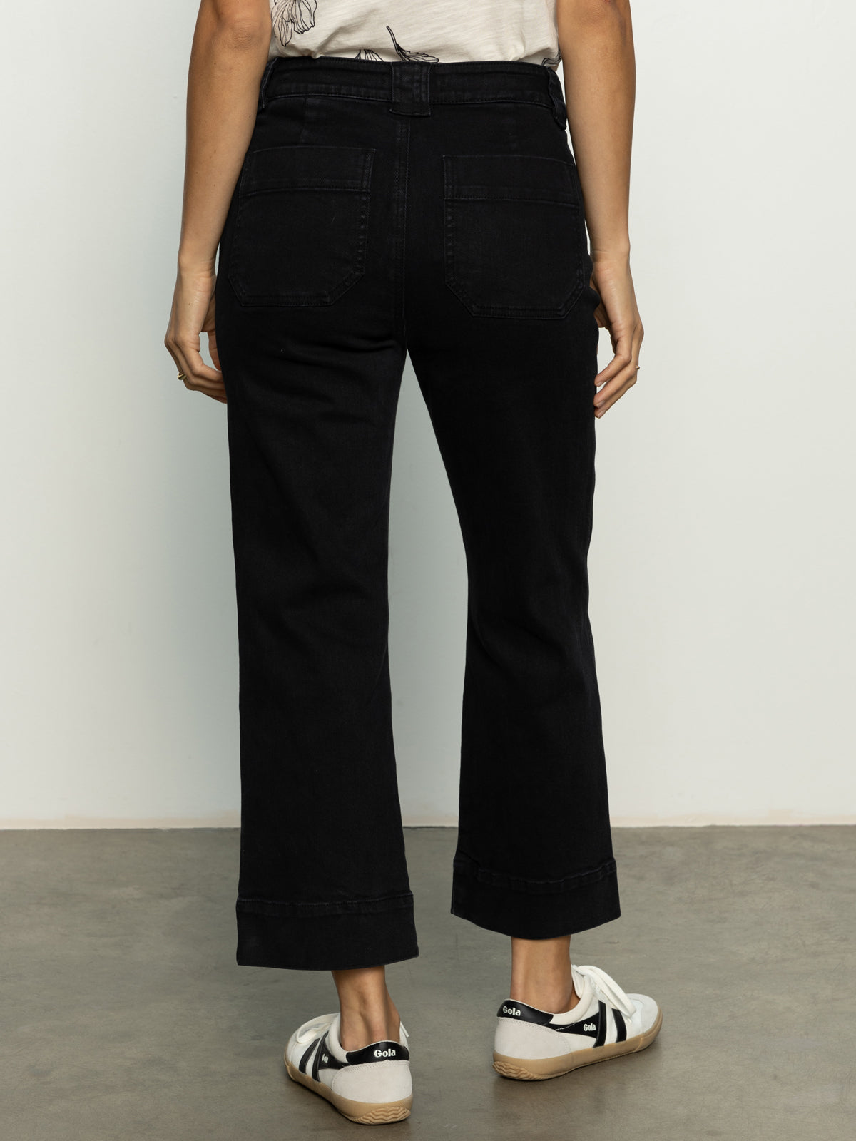 A person models Sanctuary Clothing's voyage crop pant black, featuring a high waist, back pockets, and wide legs. They wear white sneakers and stand facing away against a plain light-colored background.