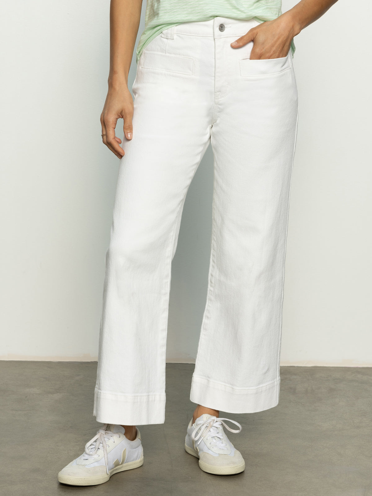 A person wearing the voyage crop pant in bleached white by Sanctuary Clothing with white sneakers stands with one hand in their pocket against a neutral-colored wall and floor.