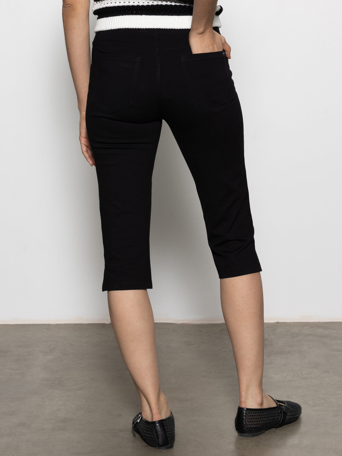 A person is wearing Sanctuary Clothing's Clipper Ponte Capri Pant in black and a black and white top, standing on a concrete floor facing a white wall. Their feet are in black woven flat-soled shoes with their arms relaxed at their sides.