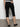 A person is wearing Sanctuary Clothing's Clipper Ponte Capri Pant in black and a black and white top, standing on a concrete floor facing a white wall. Their feet are in black woven flat-soled shoes with their arms relaxed at their sides.