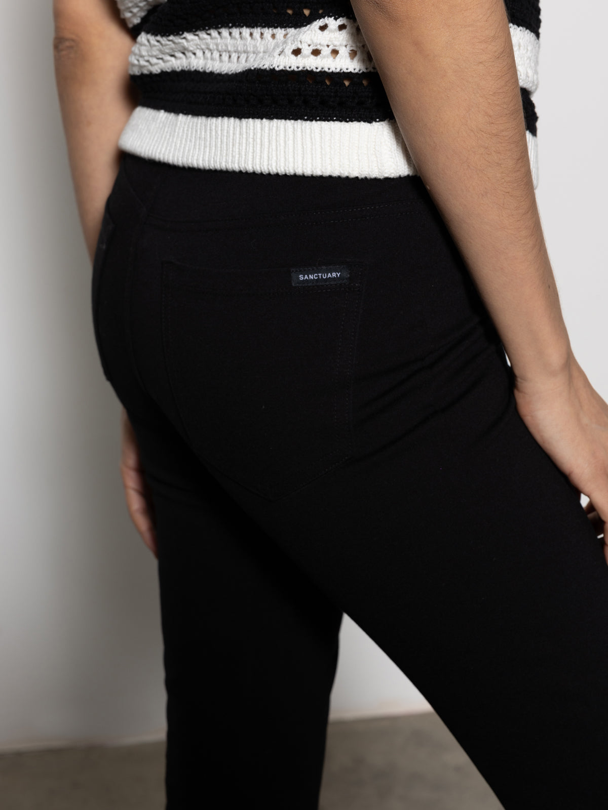 A person stands sideways wearing Sanctuary Clothing's clipper ponte capri pant in black. The focus is on the pants, showcasing a small back pocket with a label. The background remains neutral and minimalist.