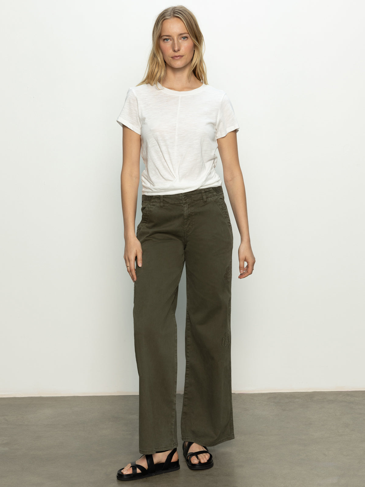 A woman stands against a plain background wearing a white T-shirt and black sandals, paired with Sanctuary Clothing's rosette army pant kalamata. Her arms are relaxed by her sides, and she is looking at the camera.