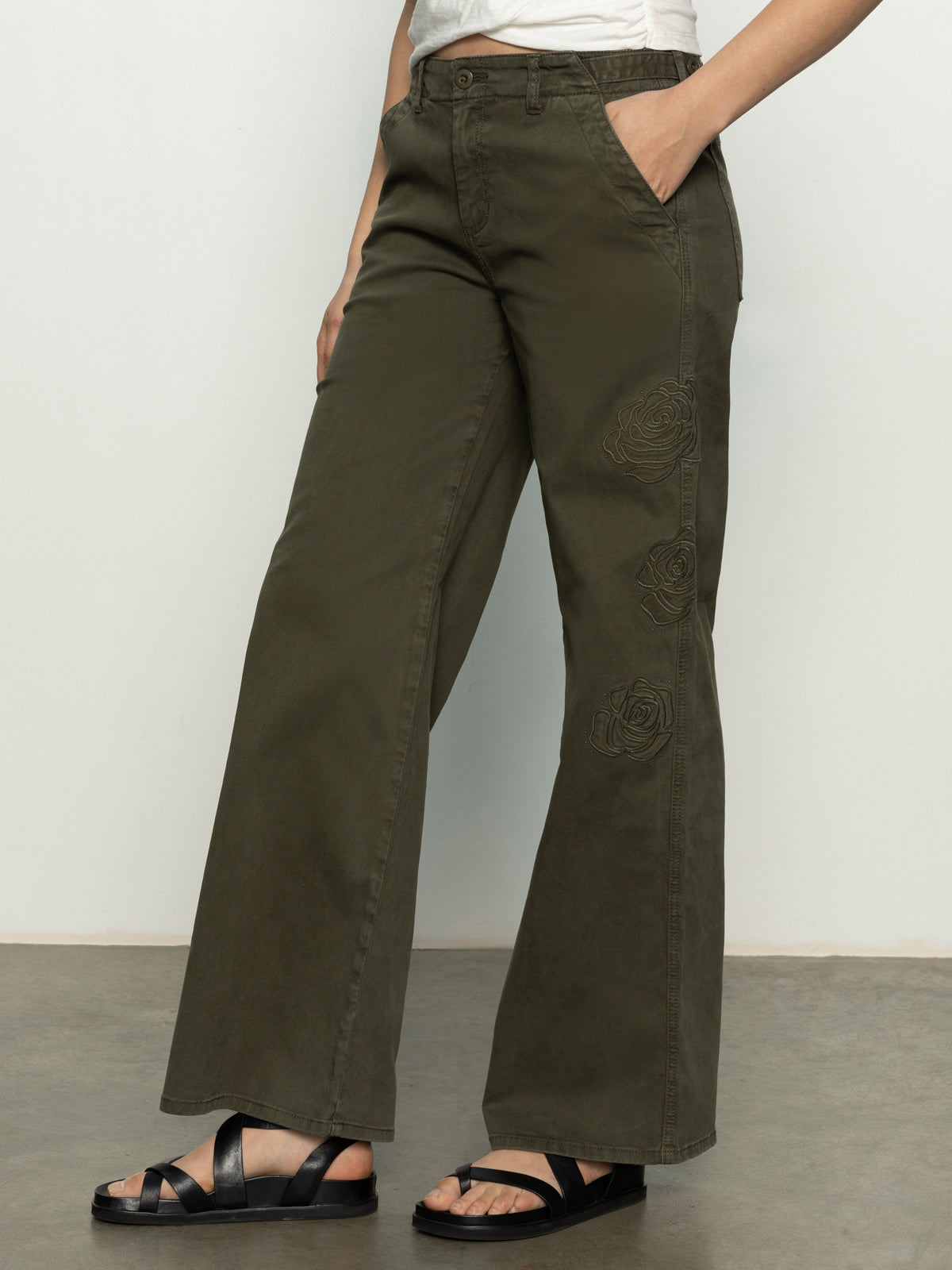 A person wearing the Sanctuary Clothing rosette army pant in kalamata, featuring floral embroidery on the side, stands against a plain background. They pair these olive green wide-leg pants with black sandals.