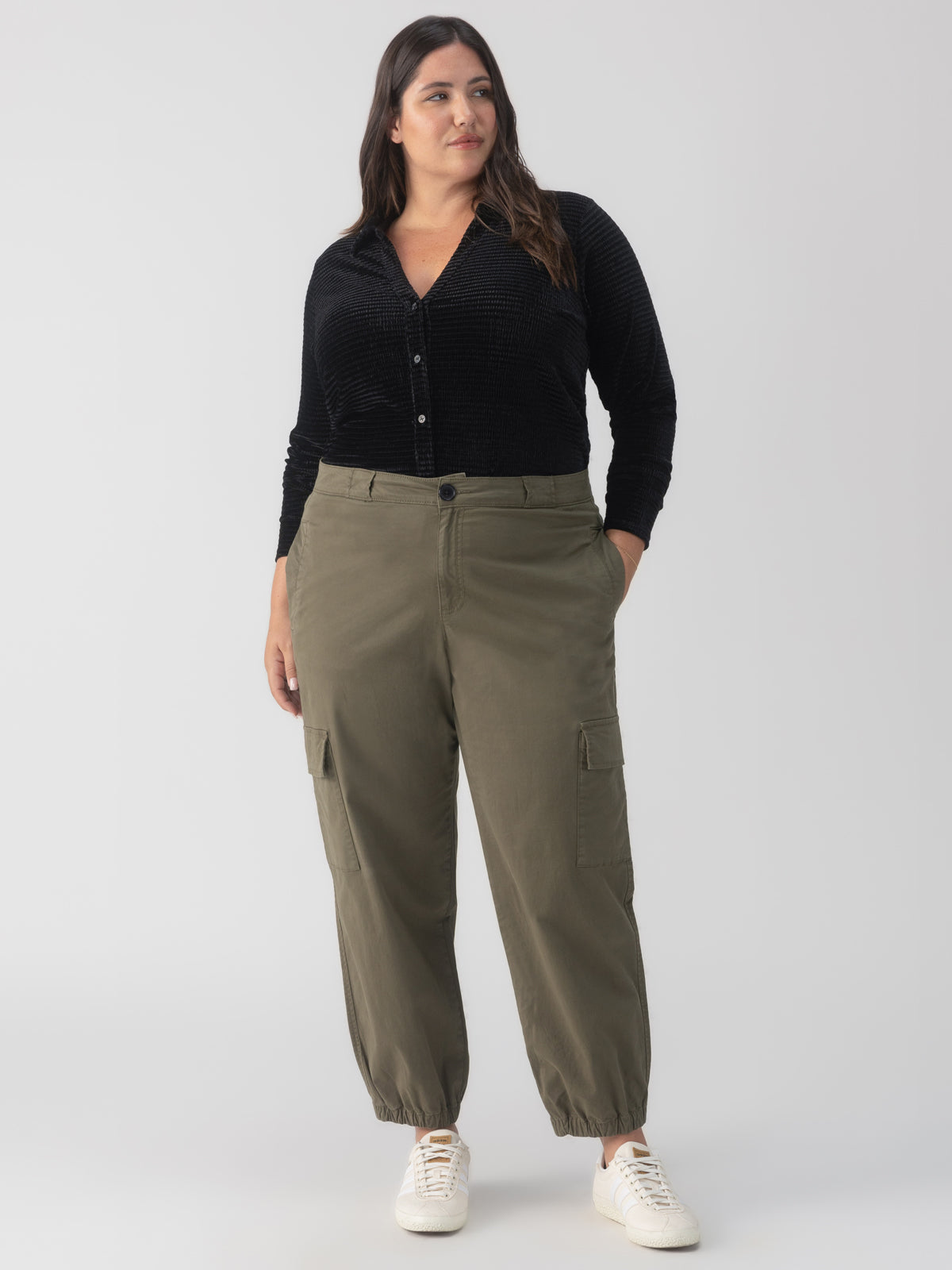 A woman is standing against a plain background. She is dressed in a black long-sleeve button-up shirt, the "Take Over Jogger Kalamata" cargo pants from the Inclusive Collection by Sanctuary Clothing, and white sneakers. Her left hand is in her pocket, and she is looking slightly to her right.