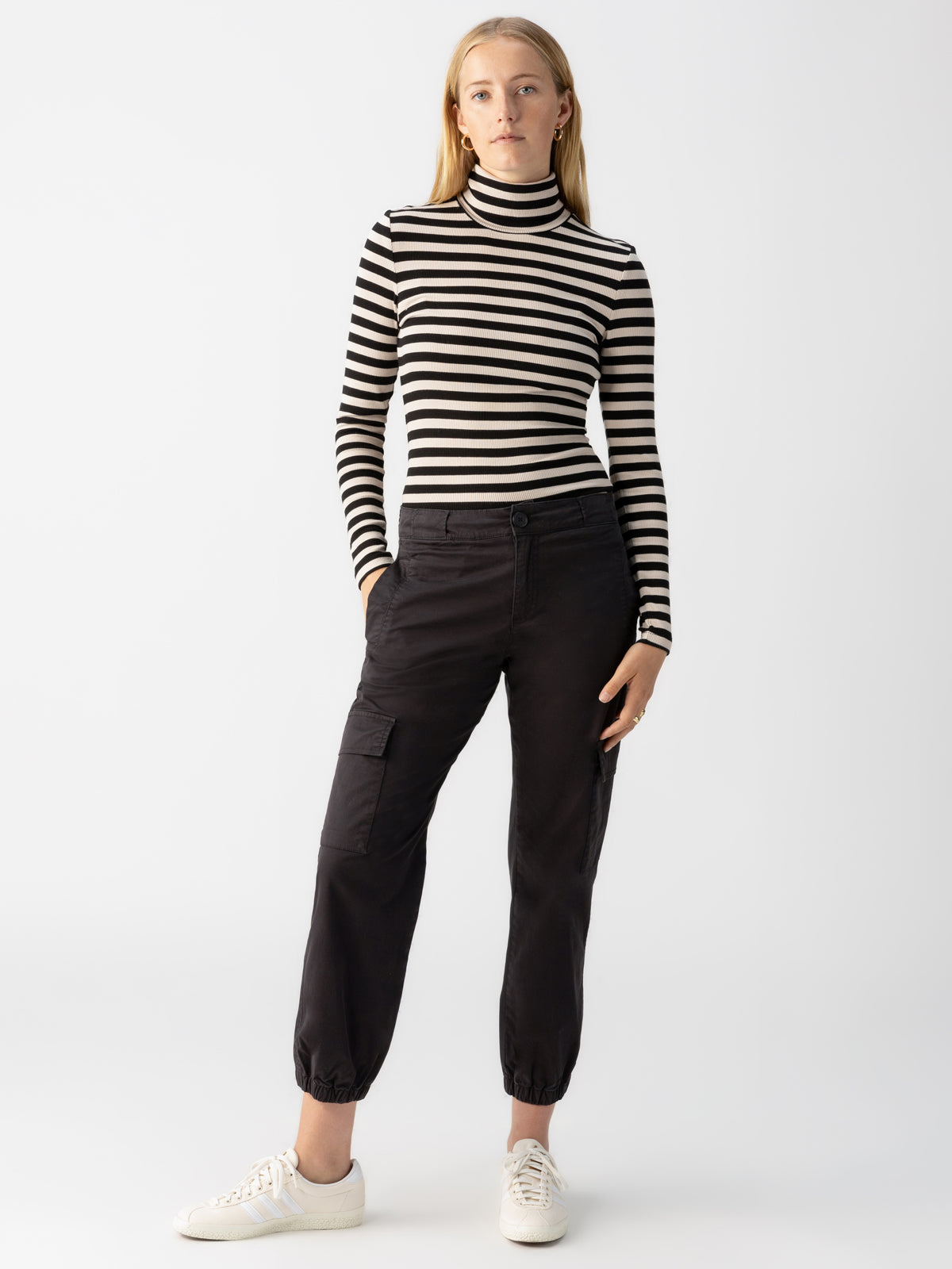 A person stands against a plain backdrop wearing a black and white striped turtleneck, Sanctuary Clothing's Take Over Jogger in black, and white sneakers. They have one hand in their pocket and a neutral expression.