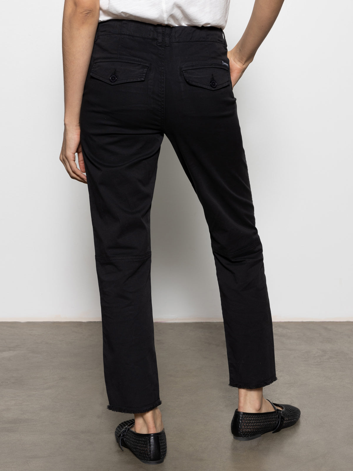 Rear view of a person wearing Sanctuary Clothing's Trade Maker Pant in black, paired with a white shirt and black woven shoes. They are standing on a gray floor against a plain white background.