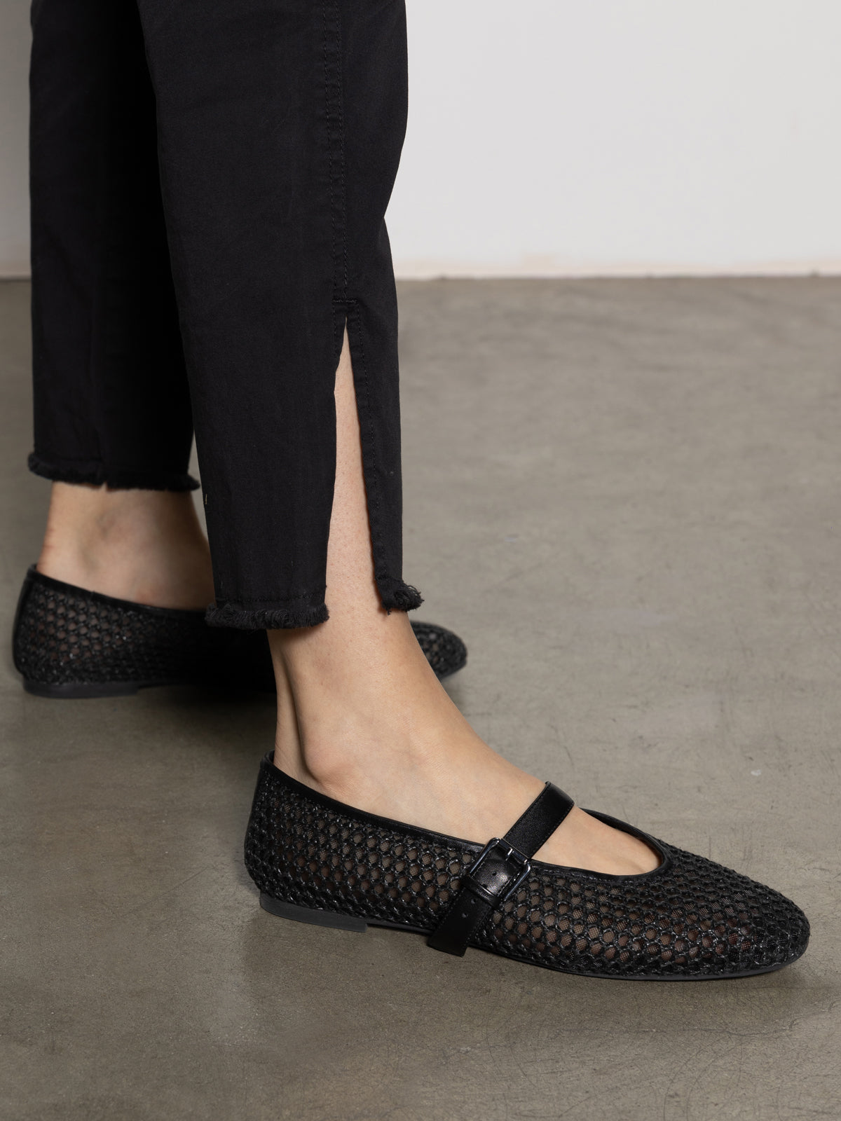 A person in Sanctuary Clothing's trade maker pant black and black woven ballet flats with a strap stands on a plain floor. The pants have a hem slit, revealing the shoes and ankle.