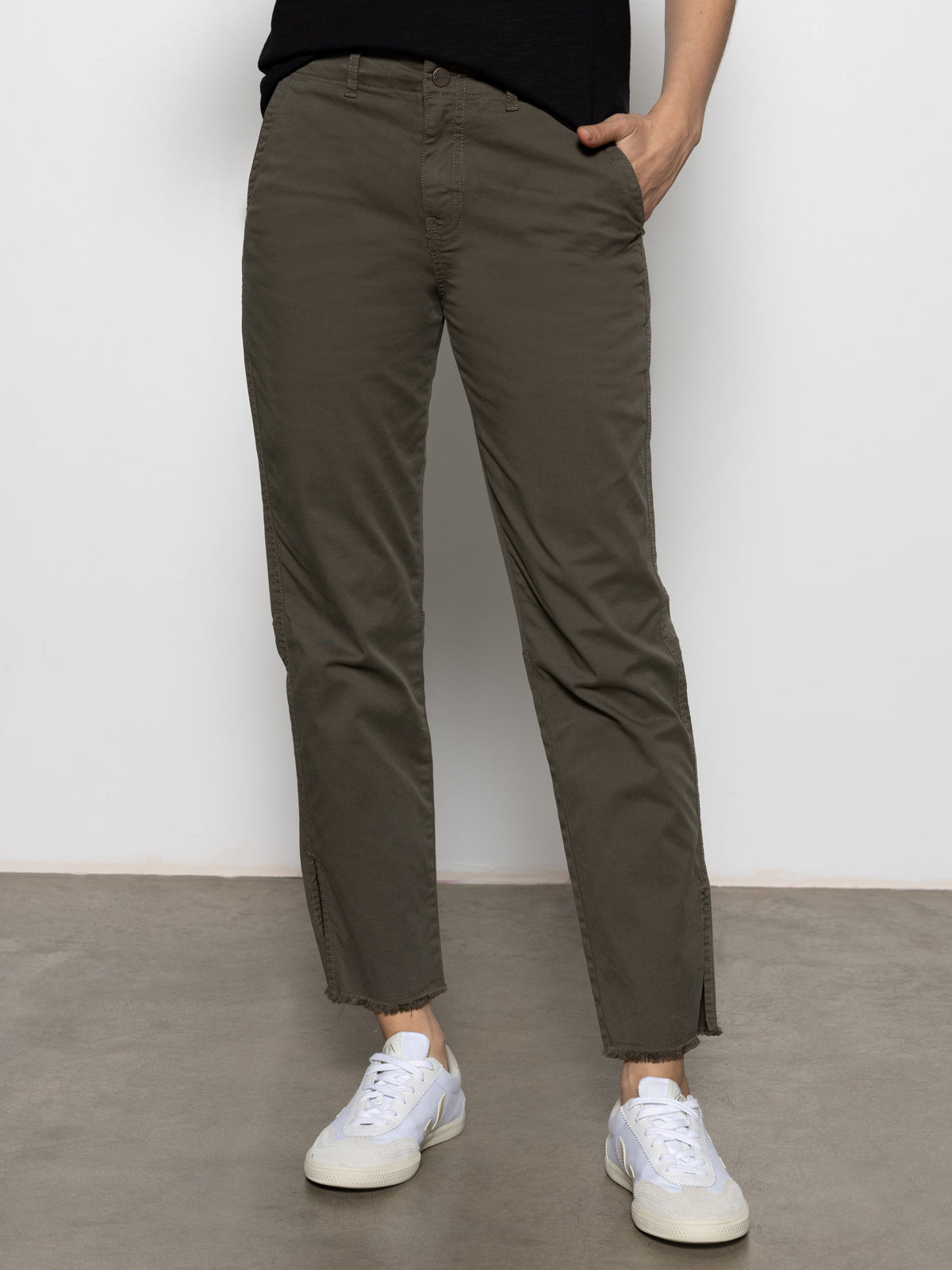 Someone wearing the Sanctuary Clothing "trade maker pant kalamata" and white sneakers stands against a plain white background. One hand is tucked into a pocket, and they are standing on a smooth gray surface. The pants have a casual, straight-leg design.